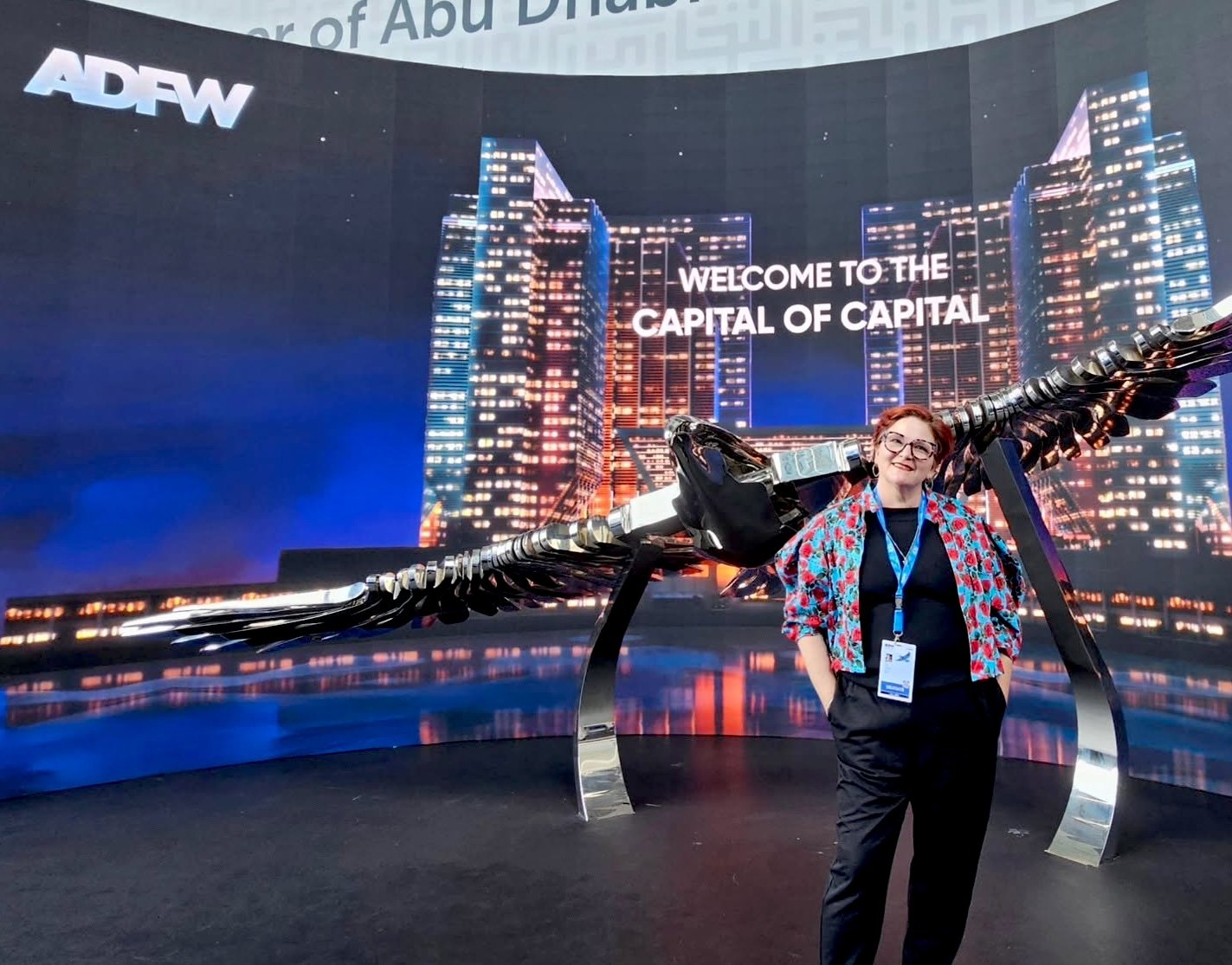 A woman standing in front of a digital backdrop with a cityscape of tall illuminated buildings. The backdrop reads "Welcome to the Capital of Capital." She is smiling, wearing glasses, a colorful floral jacket, black pants, and has an identification badge around her neck.