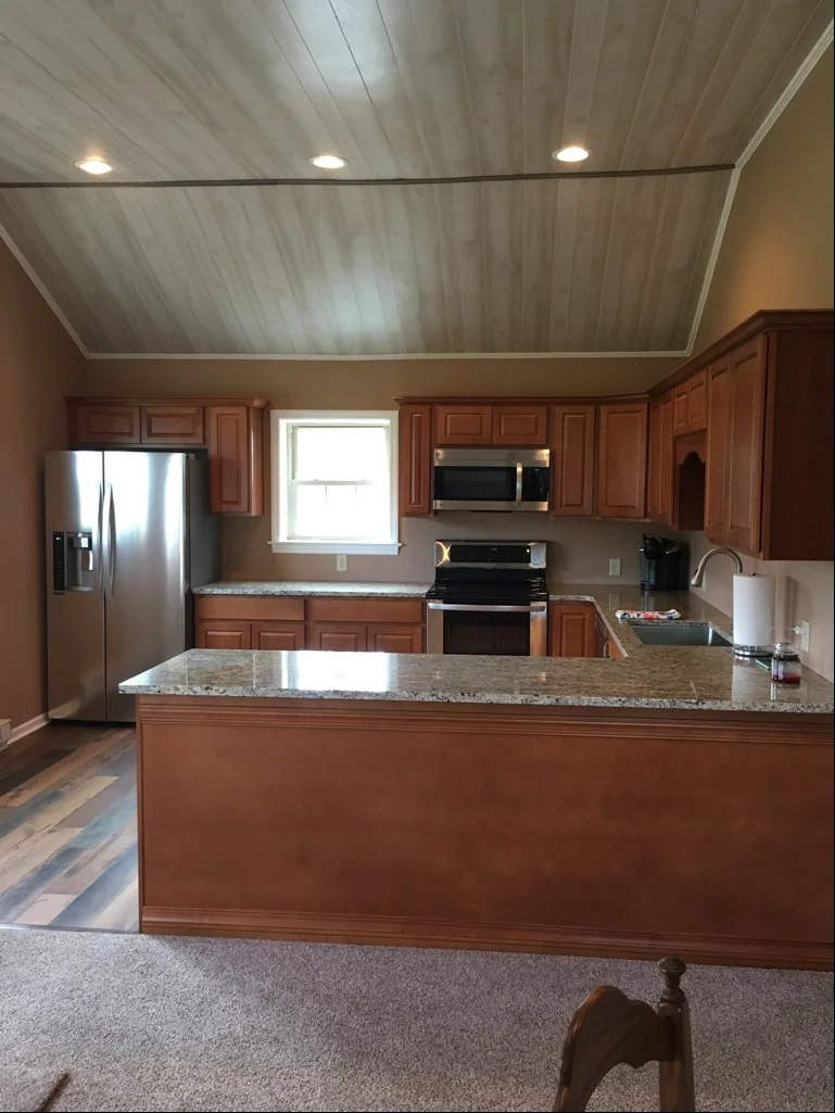 A kitchen with brown wooden cabinets, stainless steel refrigerator, microwave, and oven, granite countertops, a small window, and ceiling lights.
