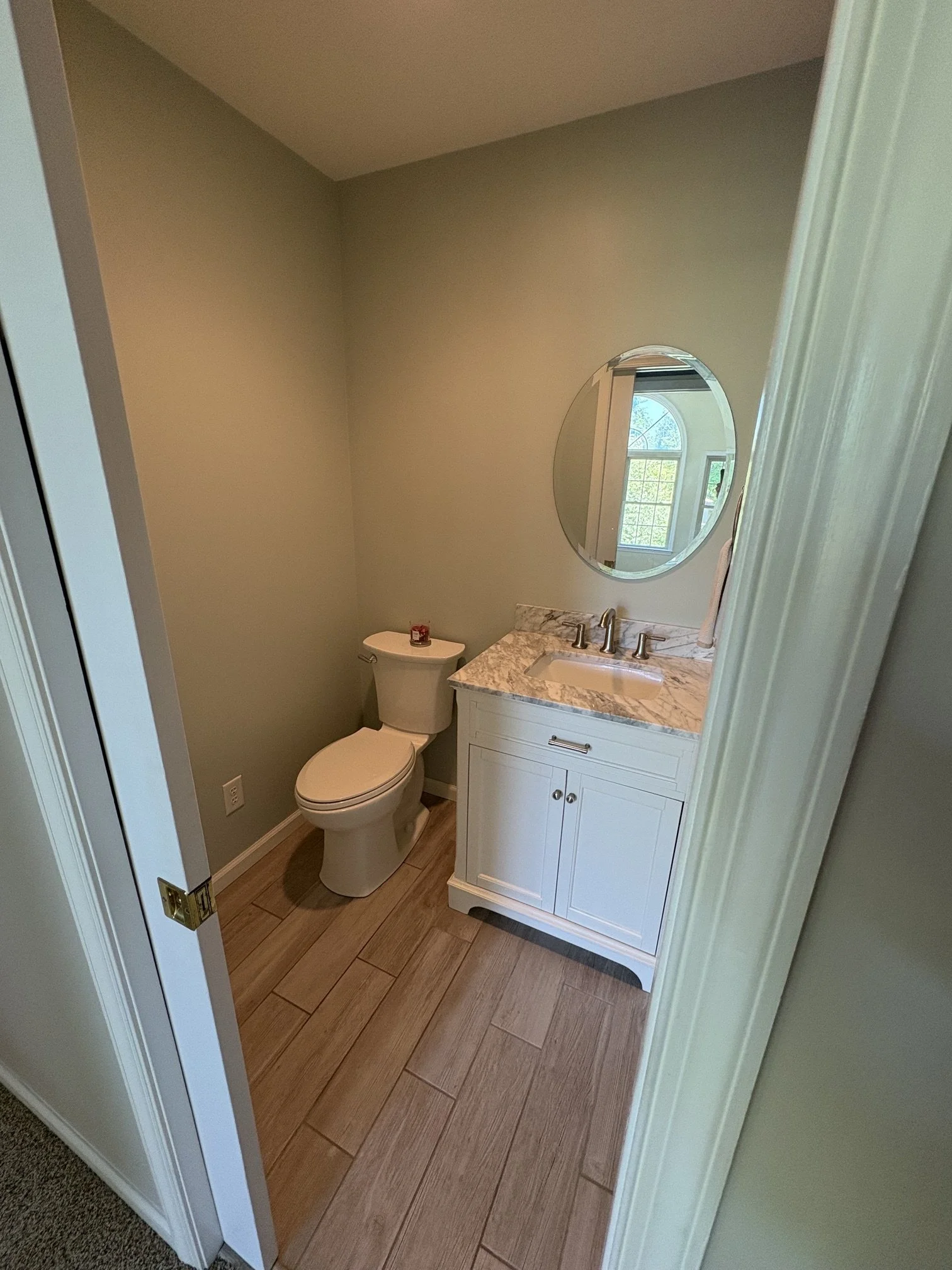 Small bathroom with beige walls, wooden flooring, a white toilet, and a white vanity with a marble countertop. There is a round mirror above the sink and a window in the reflection. A small red candle is placed on the toilet tank.