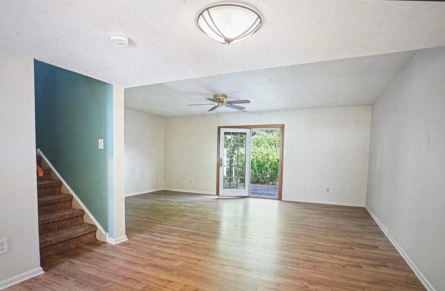 Empty living room with hardwood floors, white walls, a ceiling fan, a glass door leading outside, and a staircase with carpeted steps on the left side.