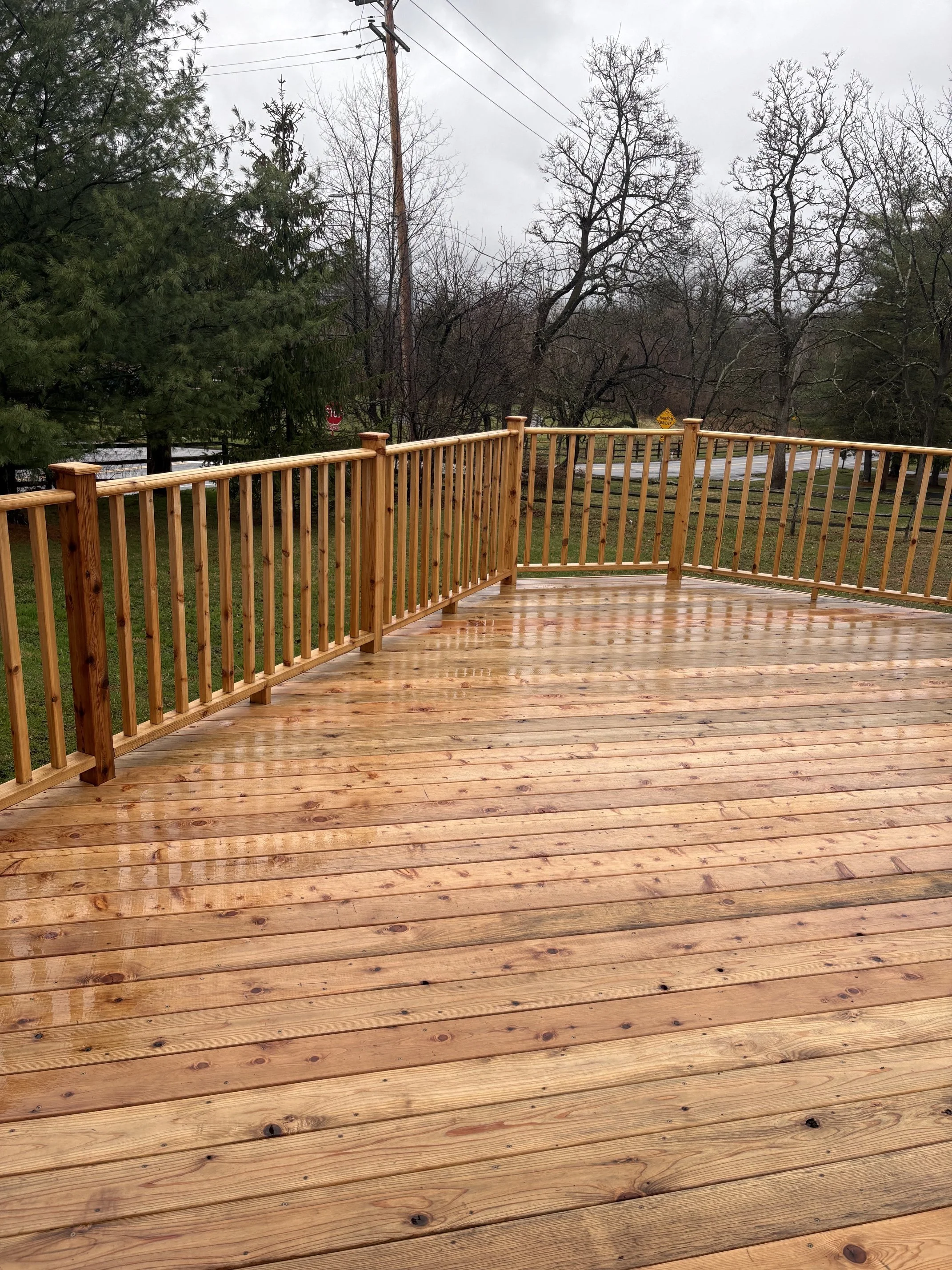 A wooden deck with a railing overlooking a grassy area with trees and power lines on a cloudy day.