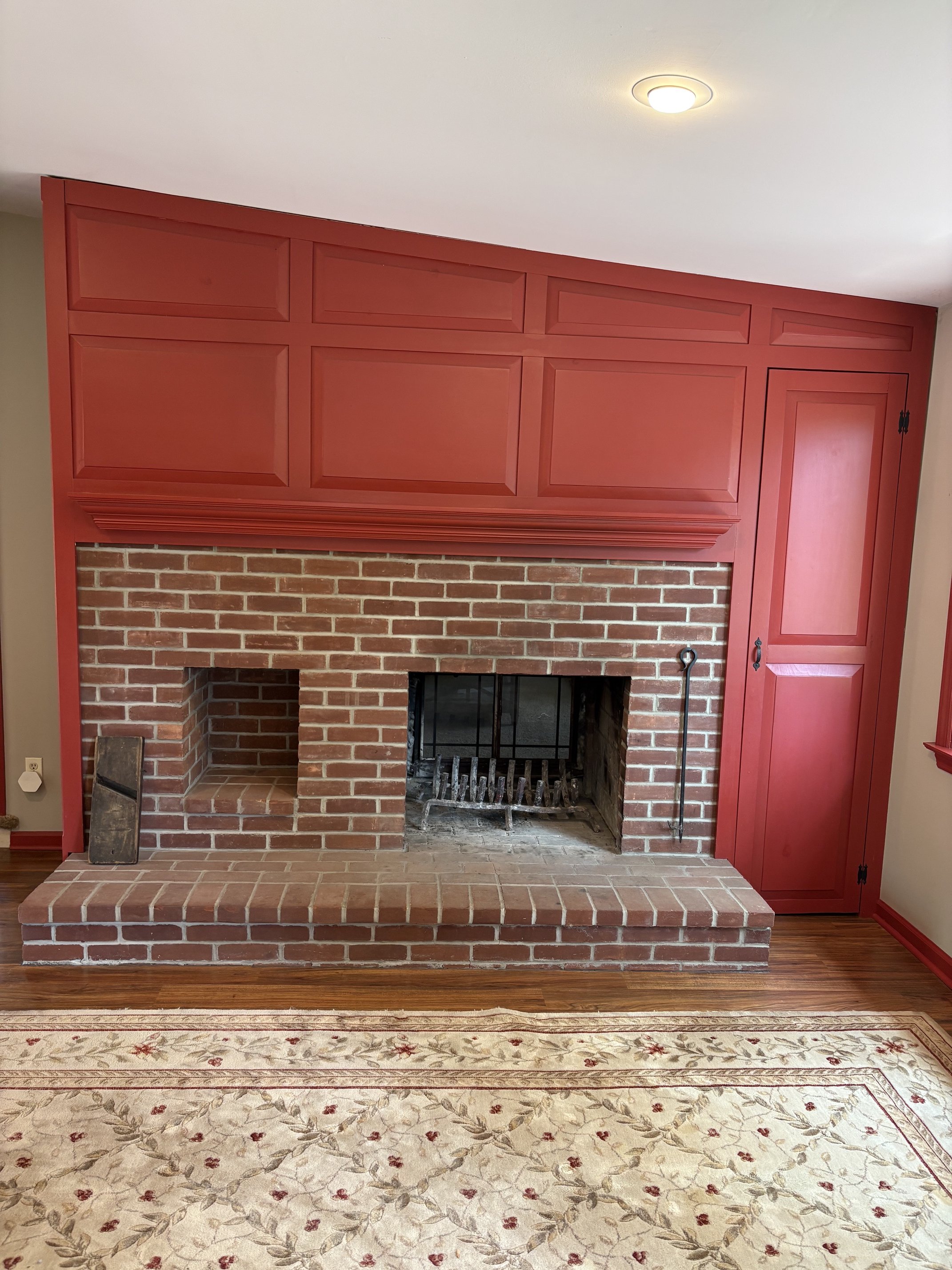 A brick fireplace with red wooden cabinetry and a mantel, with a decorative rug on a hardwood floor in front.