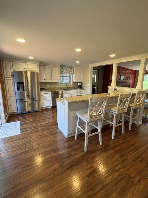 Kitchen with white cabinets, stainless steel refrigerator, granite countertops, bar seating with three chairs, hardwood floors, and recessed lighting.