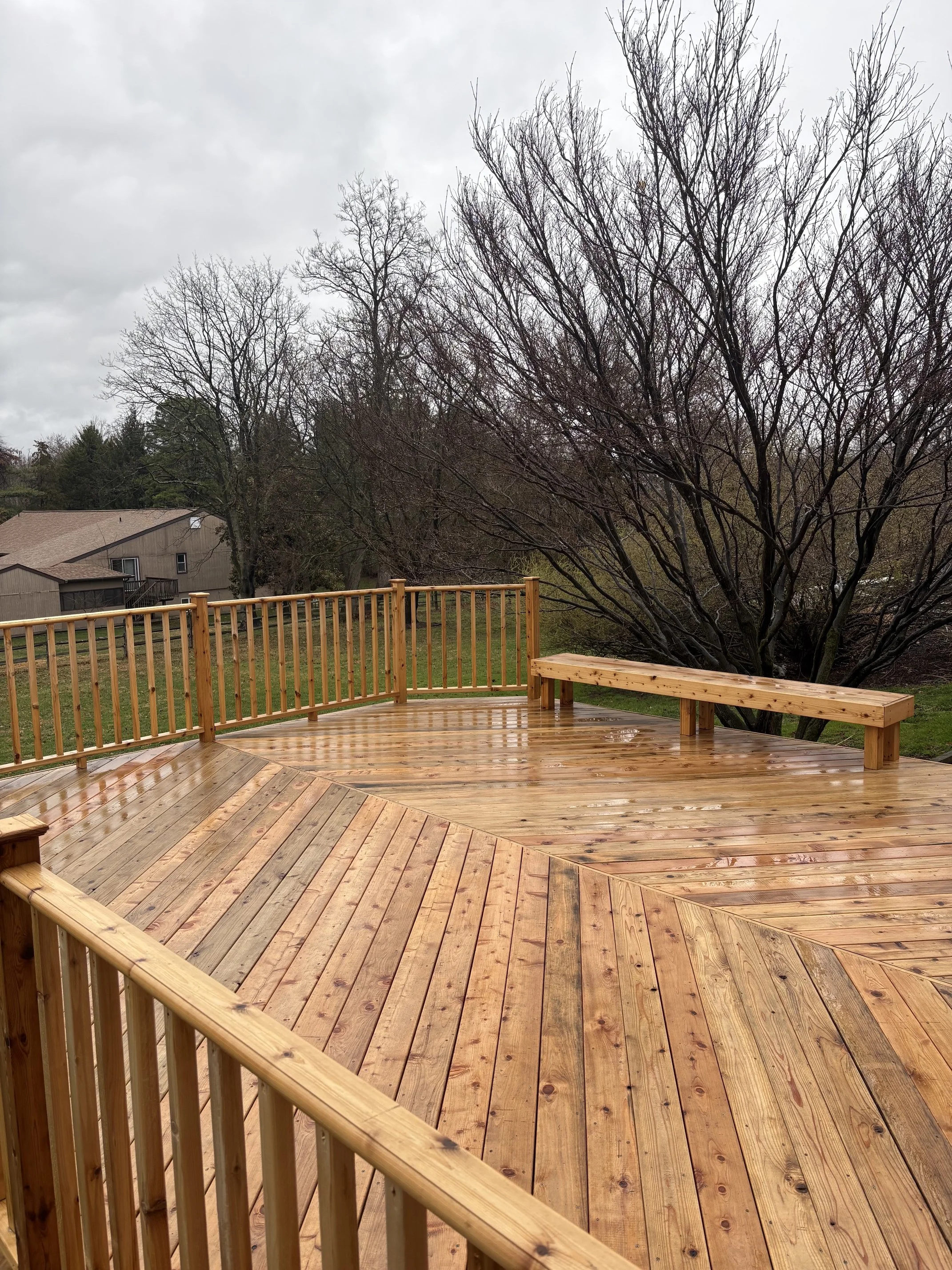 A wooden deck with a railing and a bench, overlooking a background of leafless trees and a cloudy sky.