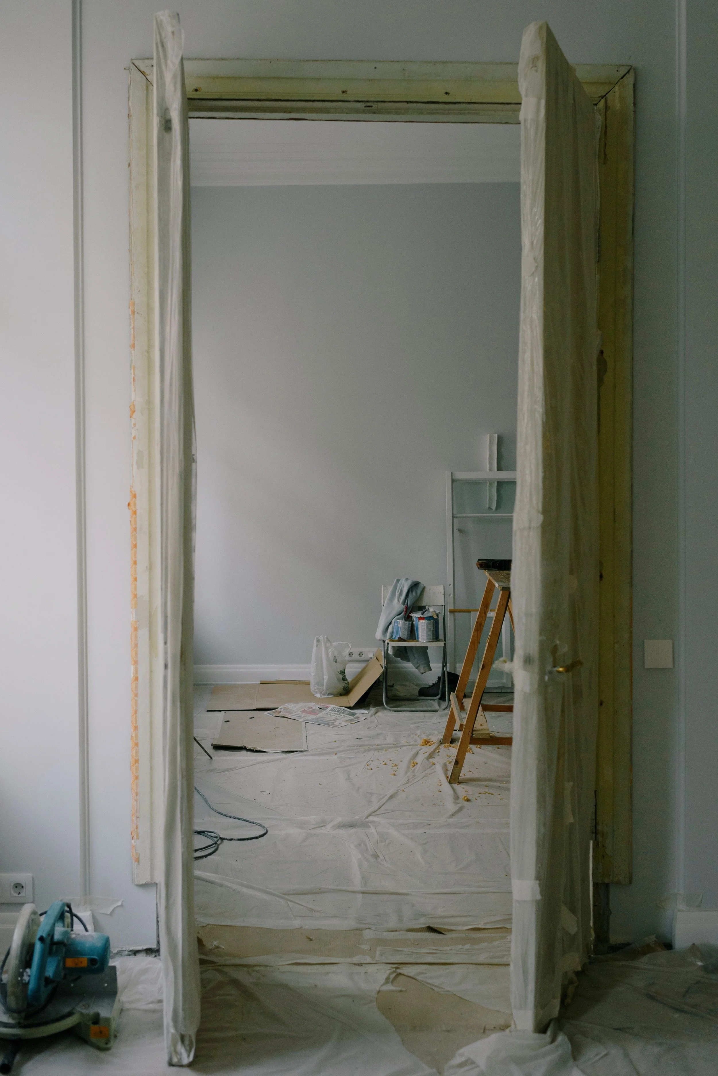 Room under renovation, viewed through doorway with partially installed door frame, construction tools and materials on the floor and a leaning ladder with supplies, paint containers, and masking tape visible in the background.