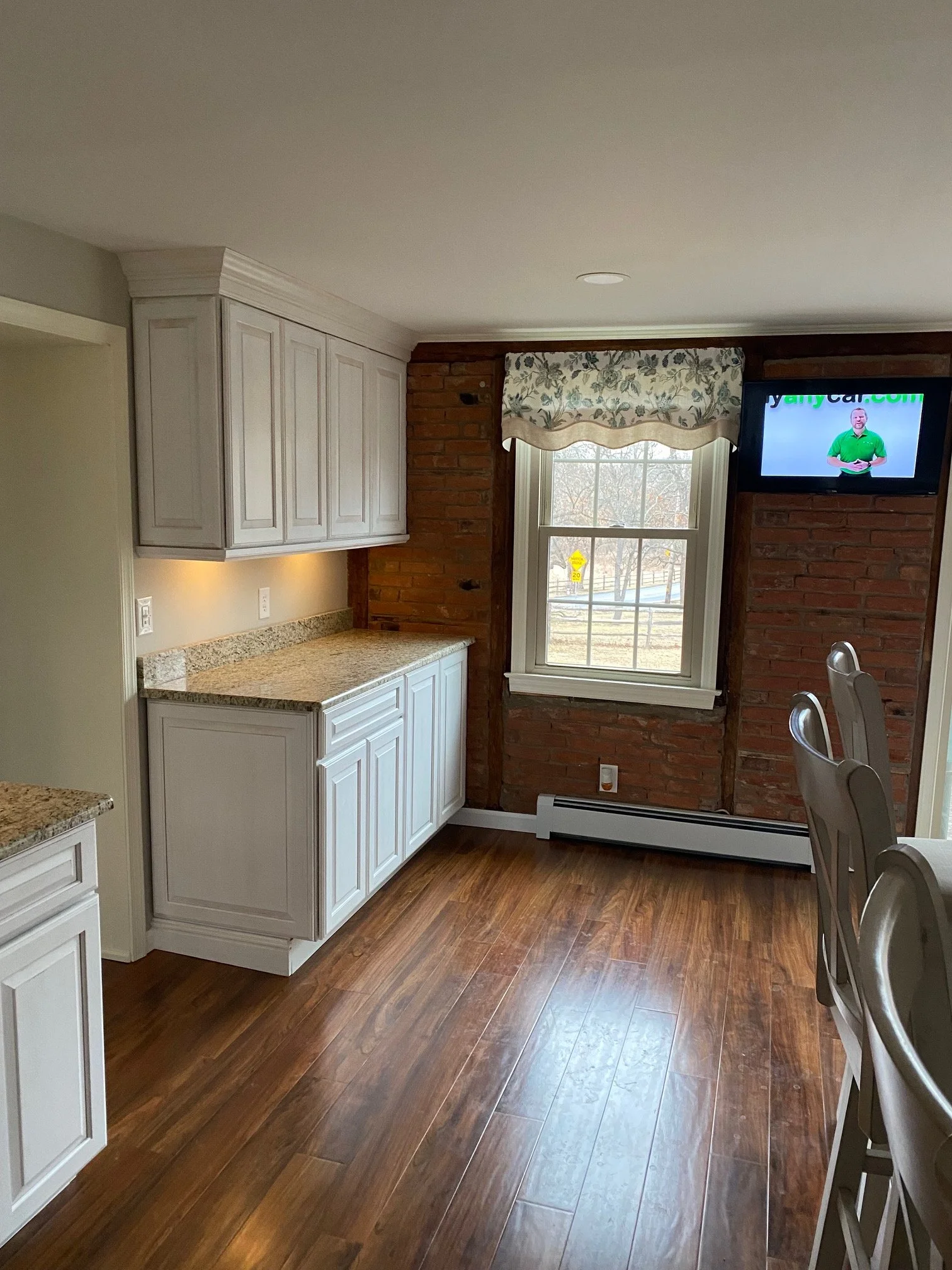Kitchen area with white cabinetry, granite countertops, a window with floral valance, and a brick wall with a mounted TV showing a man in a green shirt.