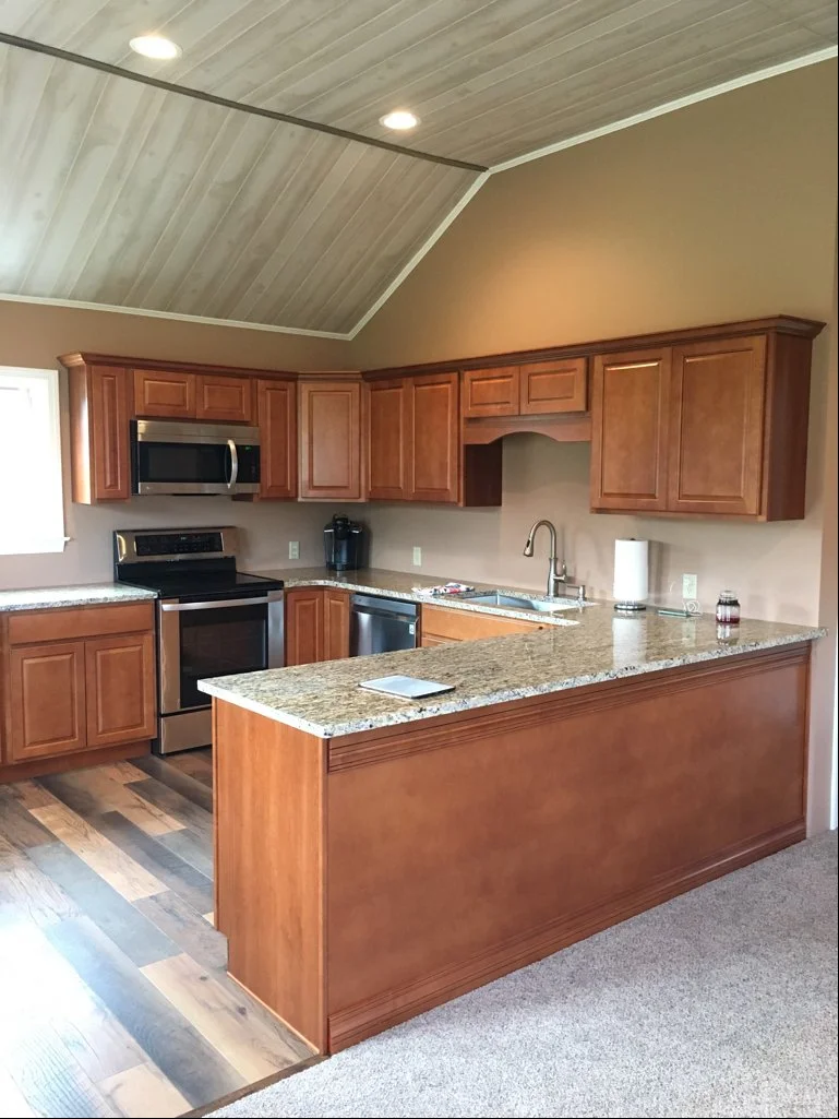 Kitchen with wooden cabinets, granite countertops, stainless steel appliances, a coffee maker, and a double-basin sink. The kitchen has a vaulted ceiling with recessed lighting and a mix of hardwood and carpeted flooring.
