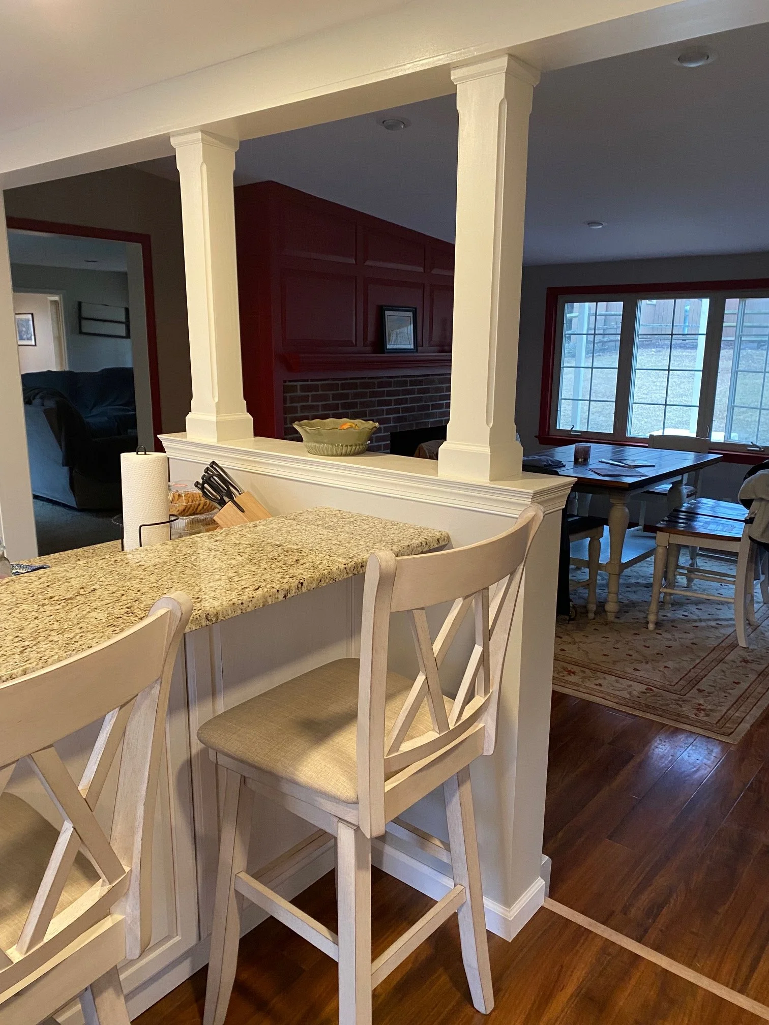 View of a kitchen counter with white wooden chairs, a paper towel holder, and a wooden handle utensil holder. The background shows part of a living area with a brick fireplace, a dining table near a large window, and a red trim around the window.