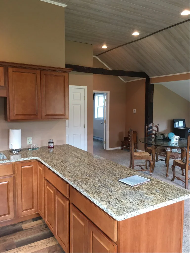 View of a kitchen with wooden cabinets, a granite countertop, and a dining area in the background with a round glass-top table and four chairs.