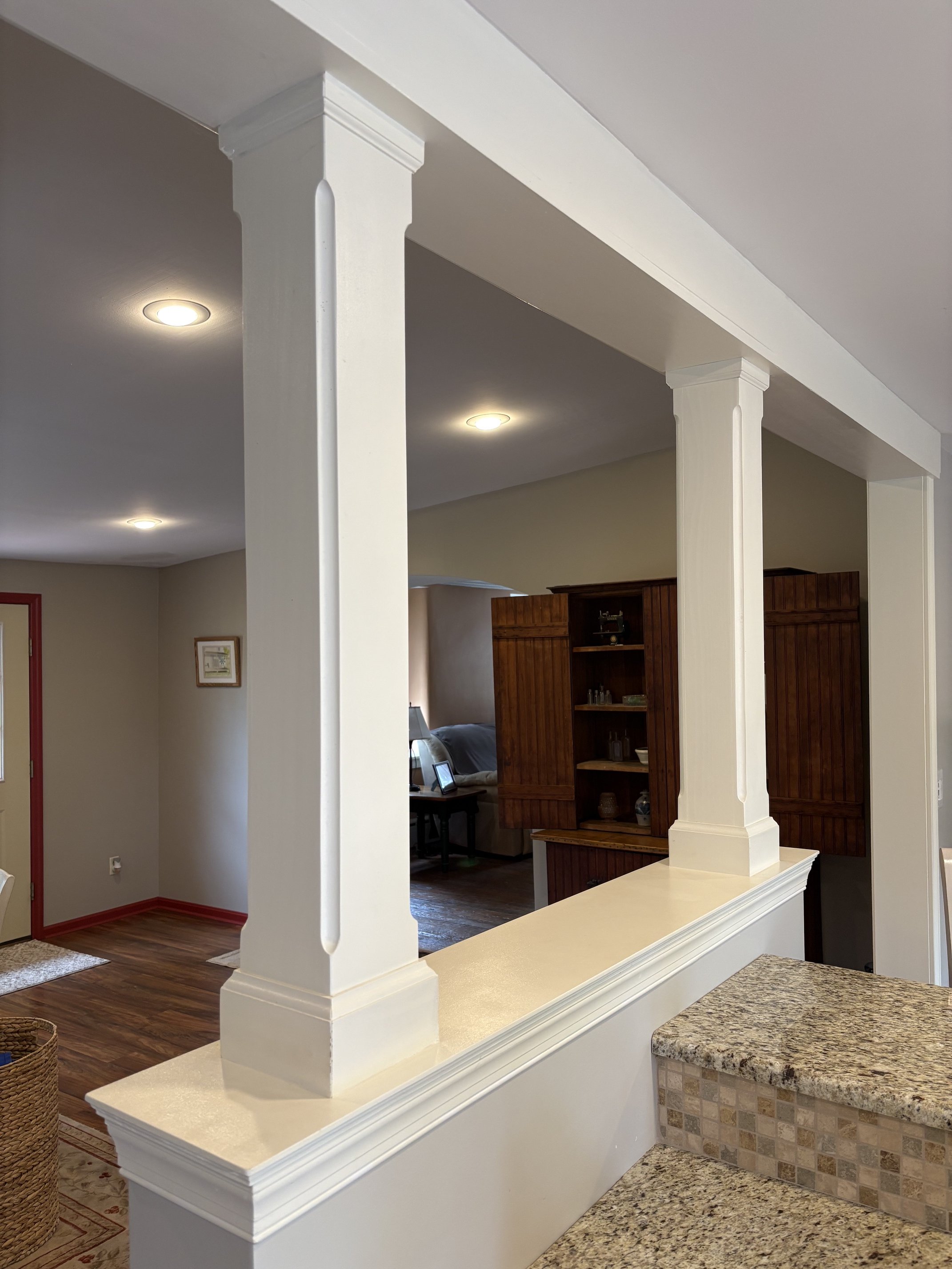 Interior view of a living space with white columns, wooden furniture, and ceiling lights, showing part of a kitchen counter and a living room with seating.