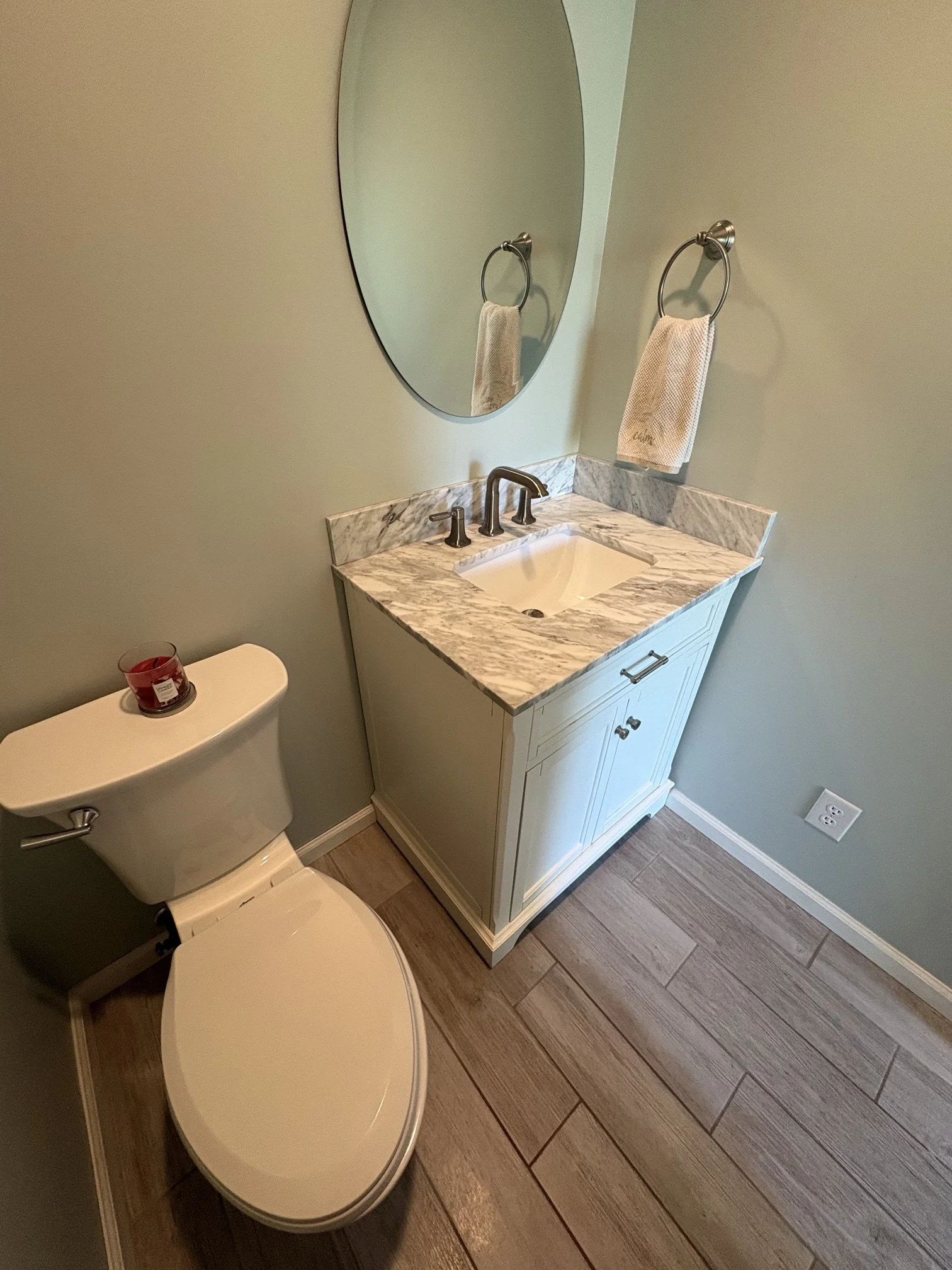 A bathroom with a toilet and a vanity with a marble top, a mirror, and a hand towel hanging on a ring. There is a candle on top of the toilet tank and a wall outlet near the floor.