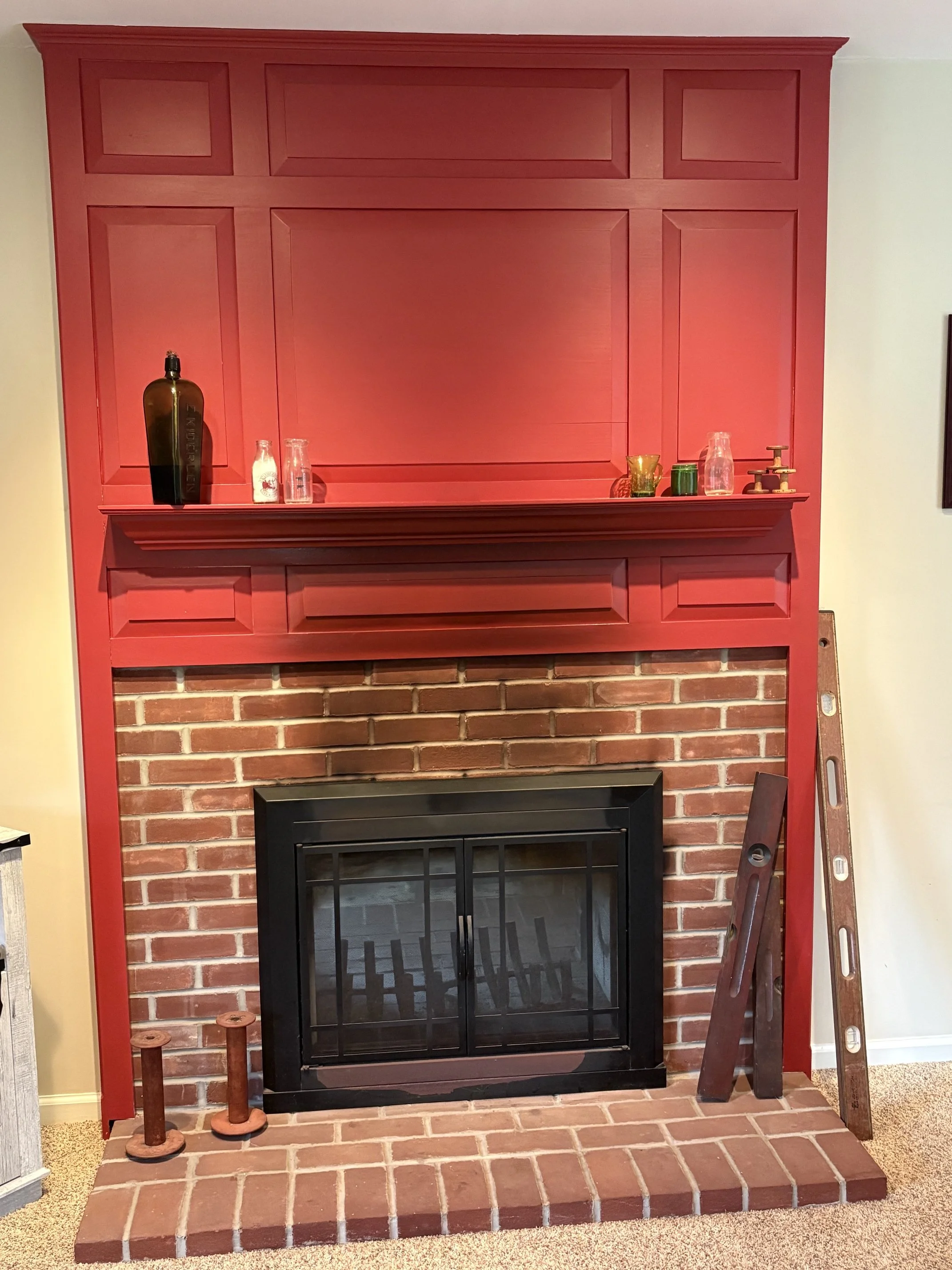 A red wooden mantel above a brick fireplace with a black metal frame. The mantel has various decorative items including bottles, glasses, and small vessels. There is a ladder leaning against the right side of the mantel. The brick hearth extends in front of the fireplace, and the surrounding wall is painted a light color.