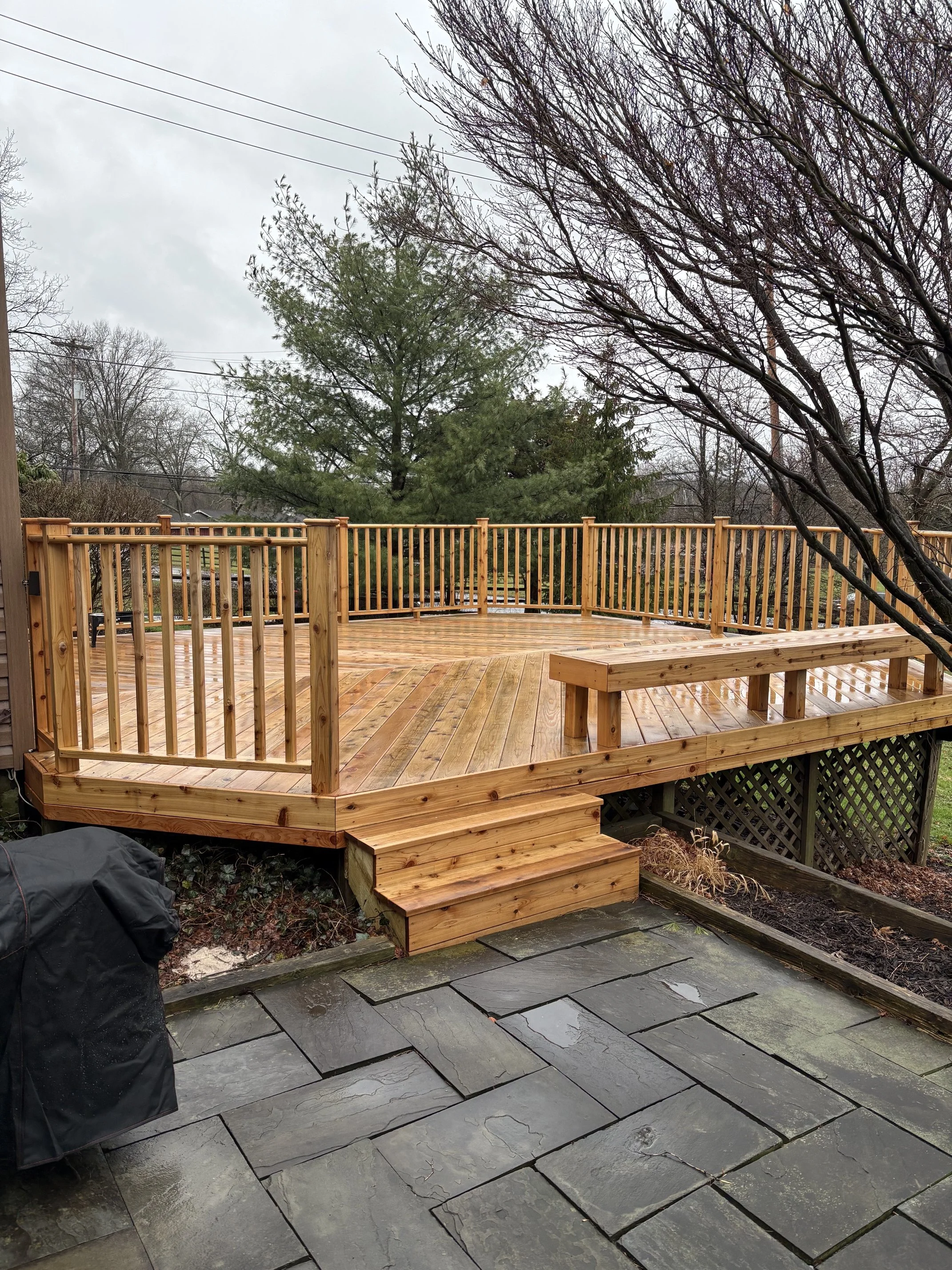 A new wooden deck with stairs and a railing in a backyard, with leafless trees and cloudy sky in the background.