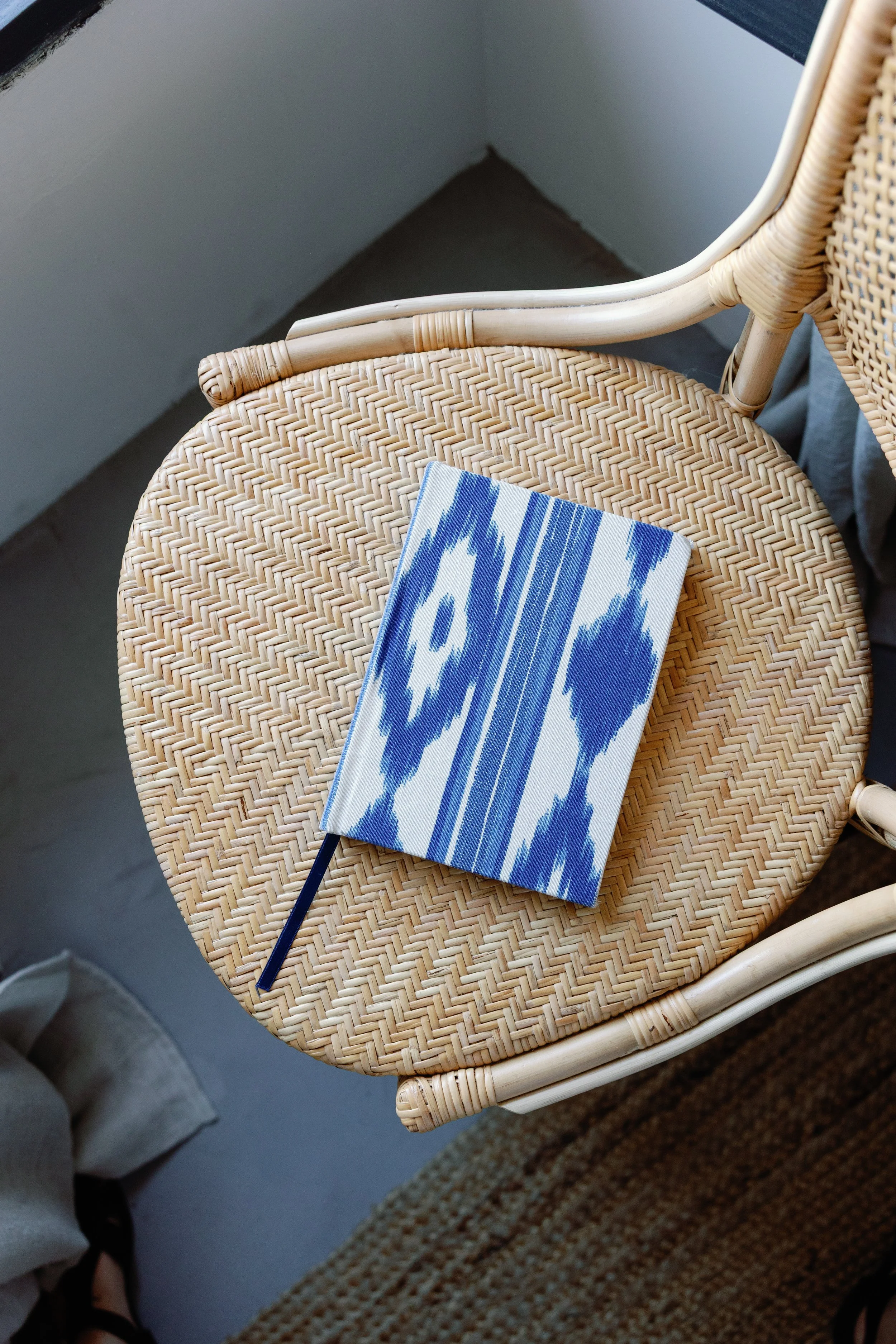 Ein Stuhl aus geflochtenem Rattan mit einem blauen und weißen Buch darauf, neben einem Regal und einem Teppich auf dem Boden.