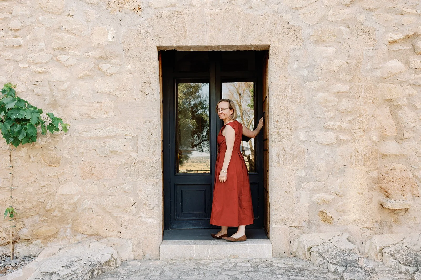 Eine Frau in rotem Kleid steht in einer schwarzen Haustür an einer Steinmauer, hält sich am Türrahmen fest und blickt nach hinten.