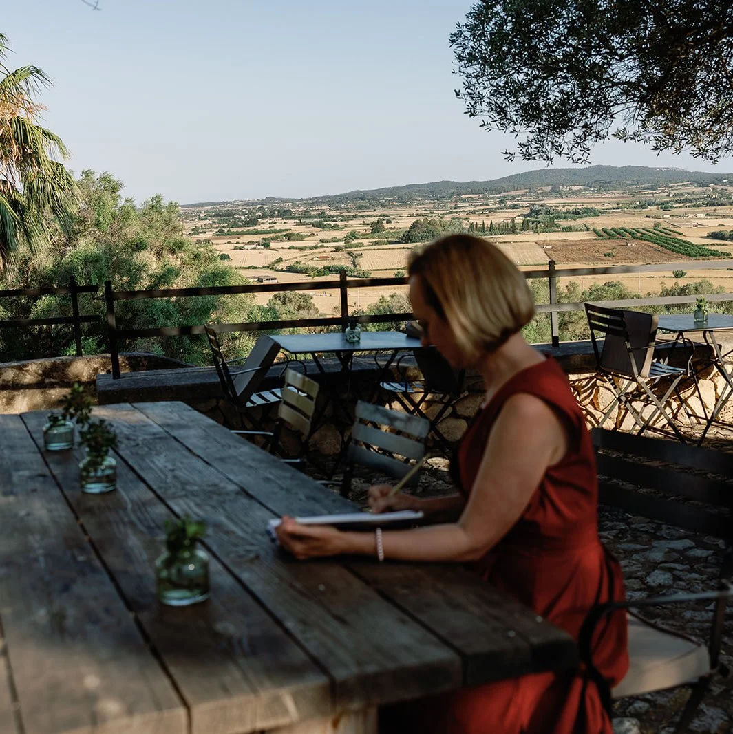 Frau in rotem Kleid sitzt an einem Holztisch im Freien mit Blick auf eine landwirtschaftliche Landschaft, während sie schreibt.