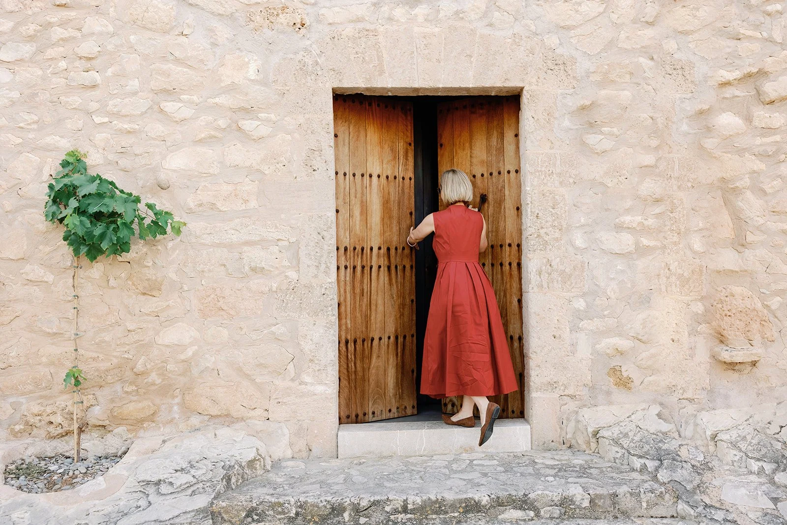 Frau in rotem Kleid öffnet eine Holzfenstertür in einer Steinmauer. Neben der Tür wächst eine Kletterpflanze.