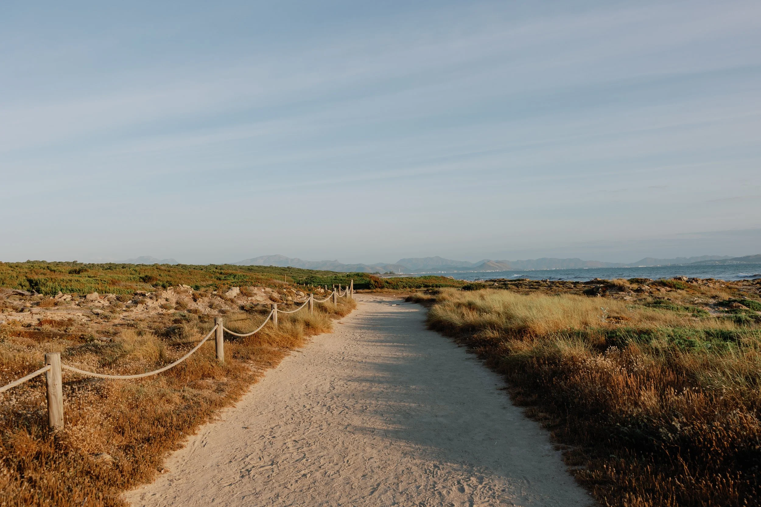 Ein Sandweg, der entlang einer Küste mit grasbewachsenen Hügeln führt, Berge im Hintergrund, Himmel mit wenigen Wolken.