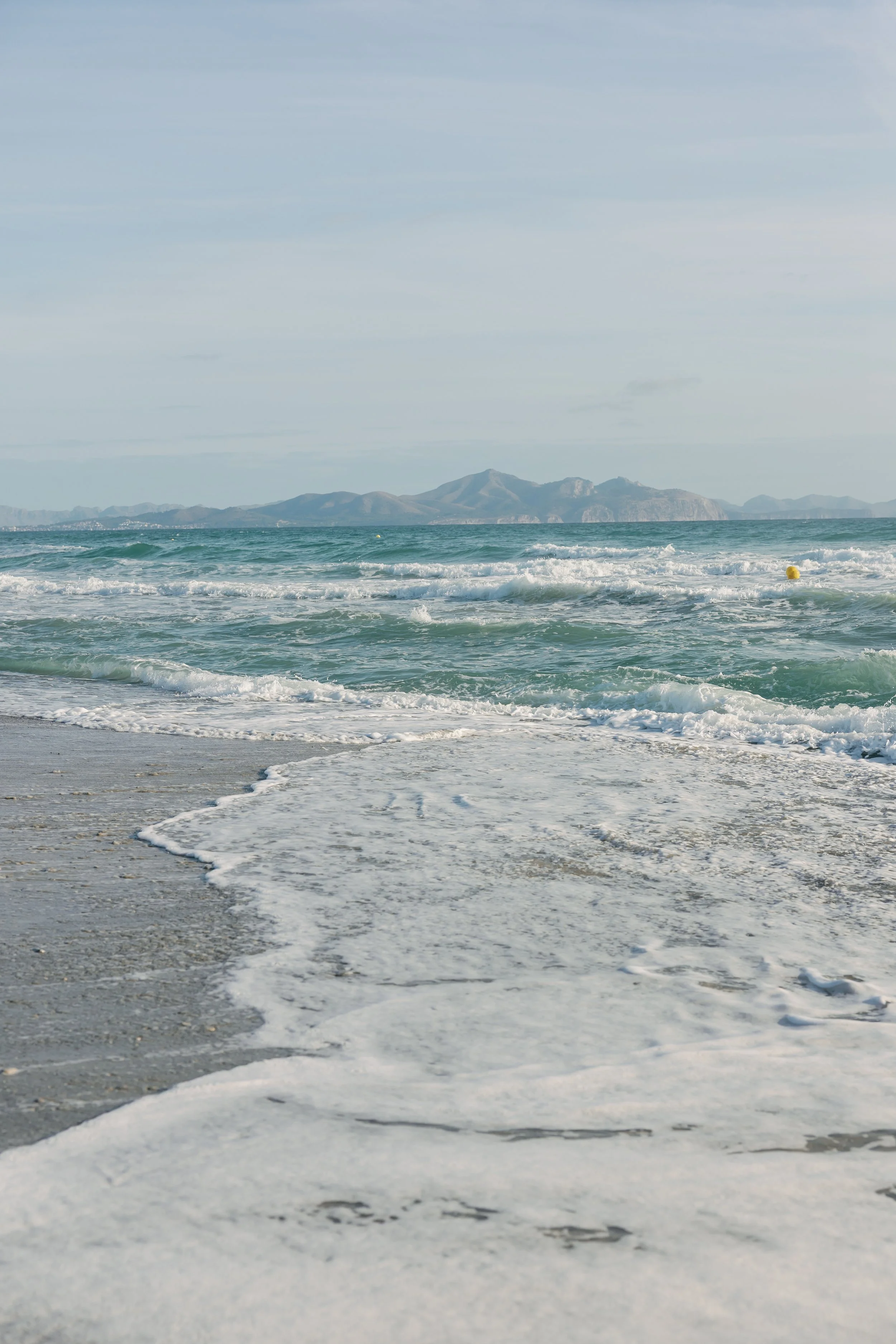 Strand mit Wellen, Sand und Bergen im Hintergrund, bewölkter Himmel