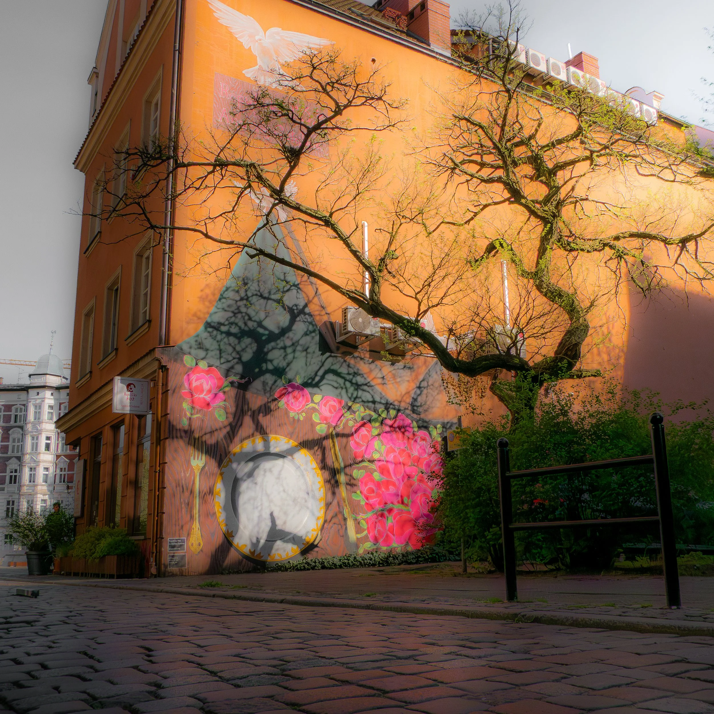 Une fresque murale représentant un oiseau blanc en vol et des fleurs roses, avec des arbres dénudés en premier plan, située sur le côté d'un bâtiment orange en ville.