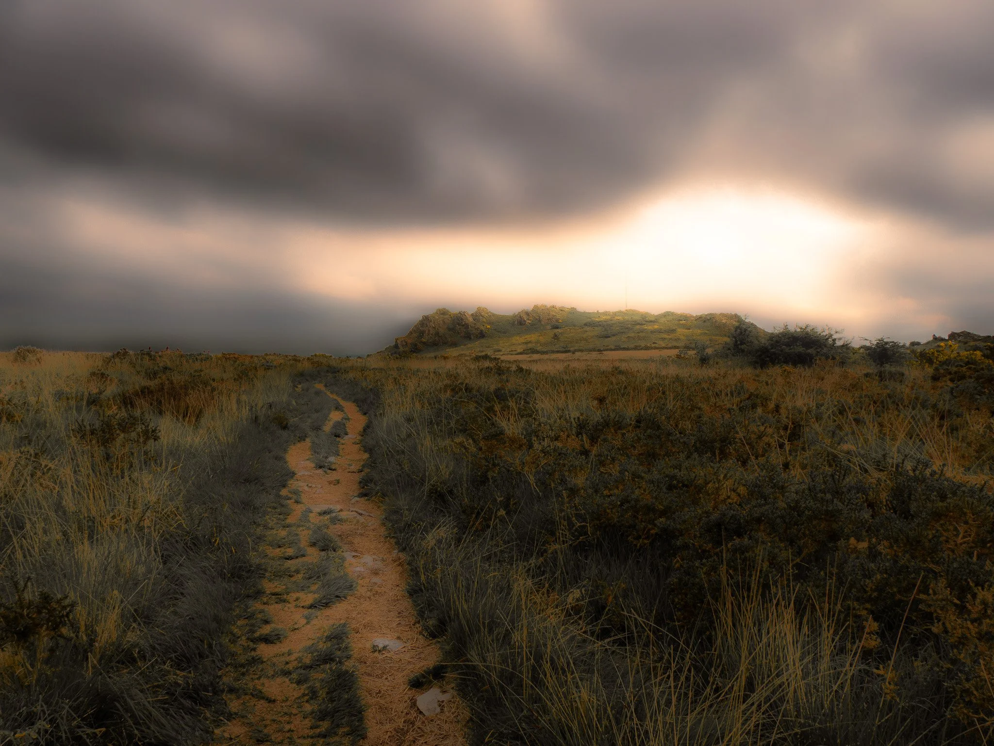 Une route de terre serpentant à travers un paysage de prairies avec des collines verdoyantes sous un ciel nuageux, avec une lumière douce de coucher de soleil.