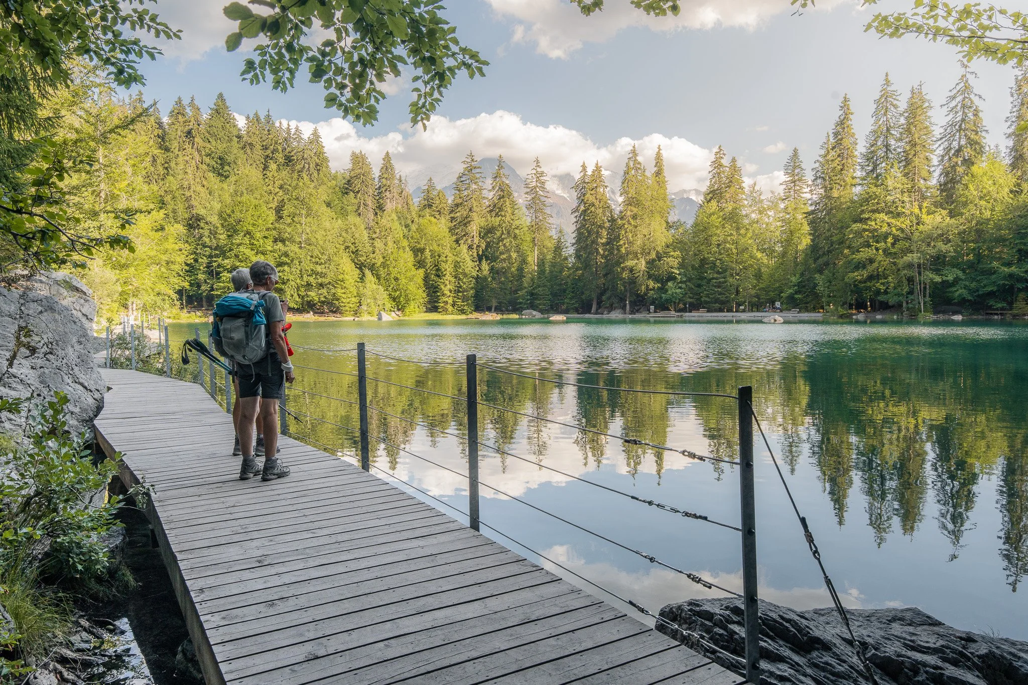 Deux personnes avec un sac à dos regardant un lac entouré de forêts de conifères en montagne, sous un ciel partiellement nuageux.