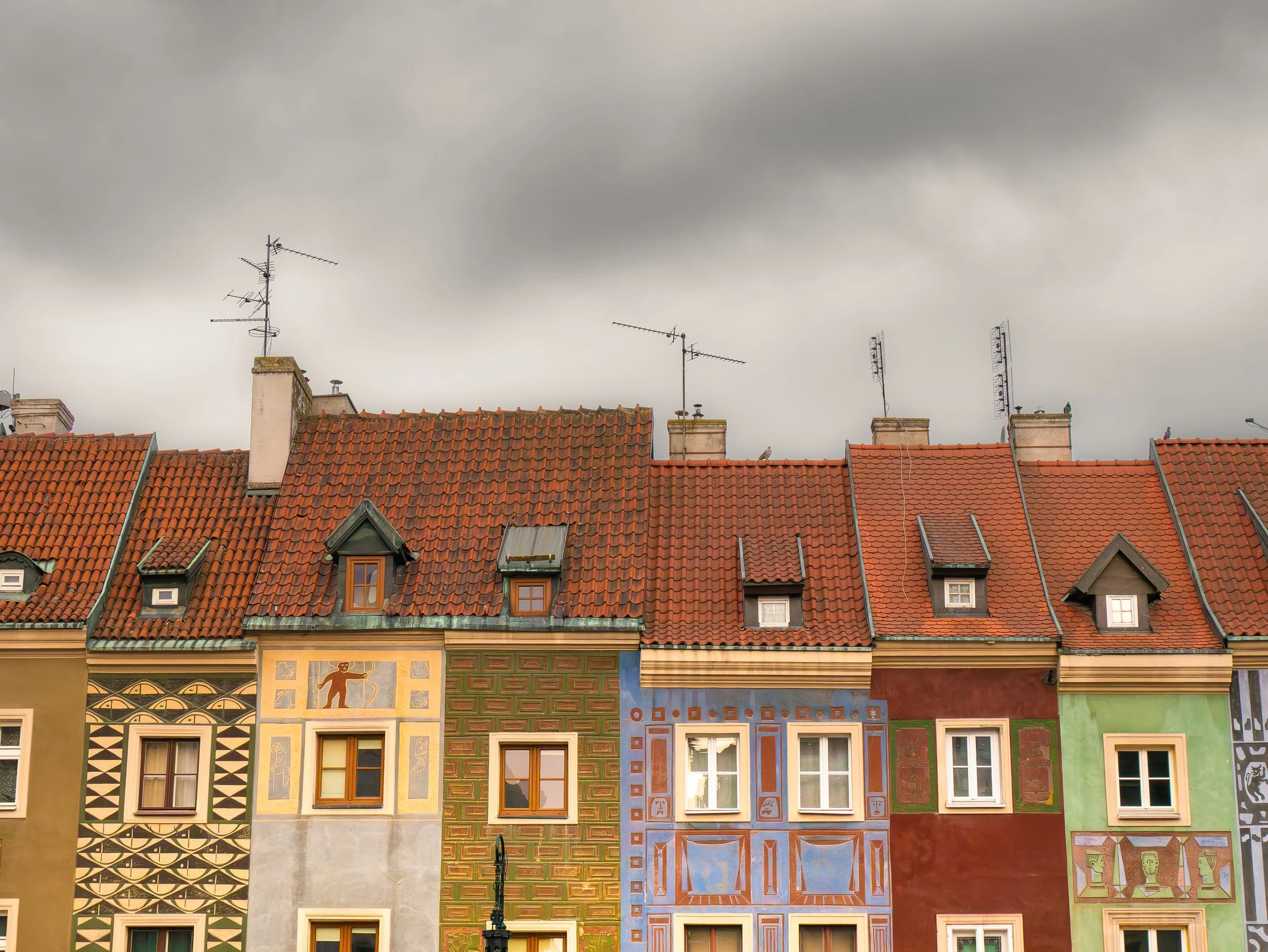Façade colorée de bâtiments avec des murs peints de motifs géométriques et de figures, fenêtres en bois, toits en tuiles rouges, et plusieurs antennes télévisées sur le toit, sous un ciel nuageux.