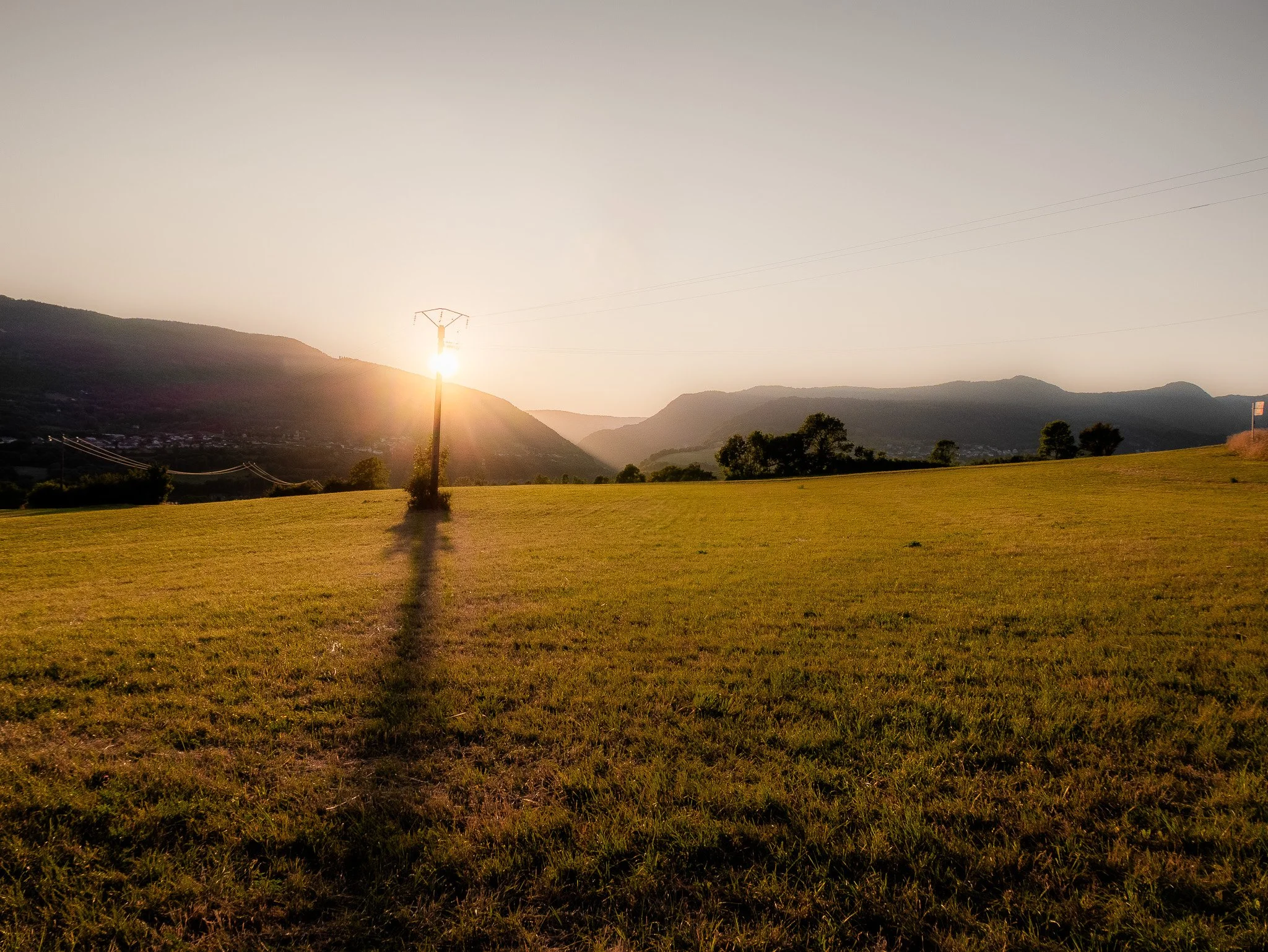 Paysage avec des montagnes, un champ vert, un pylône électrique et un soleil couchant à l'horizon.