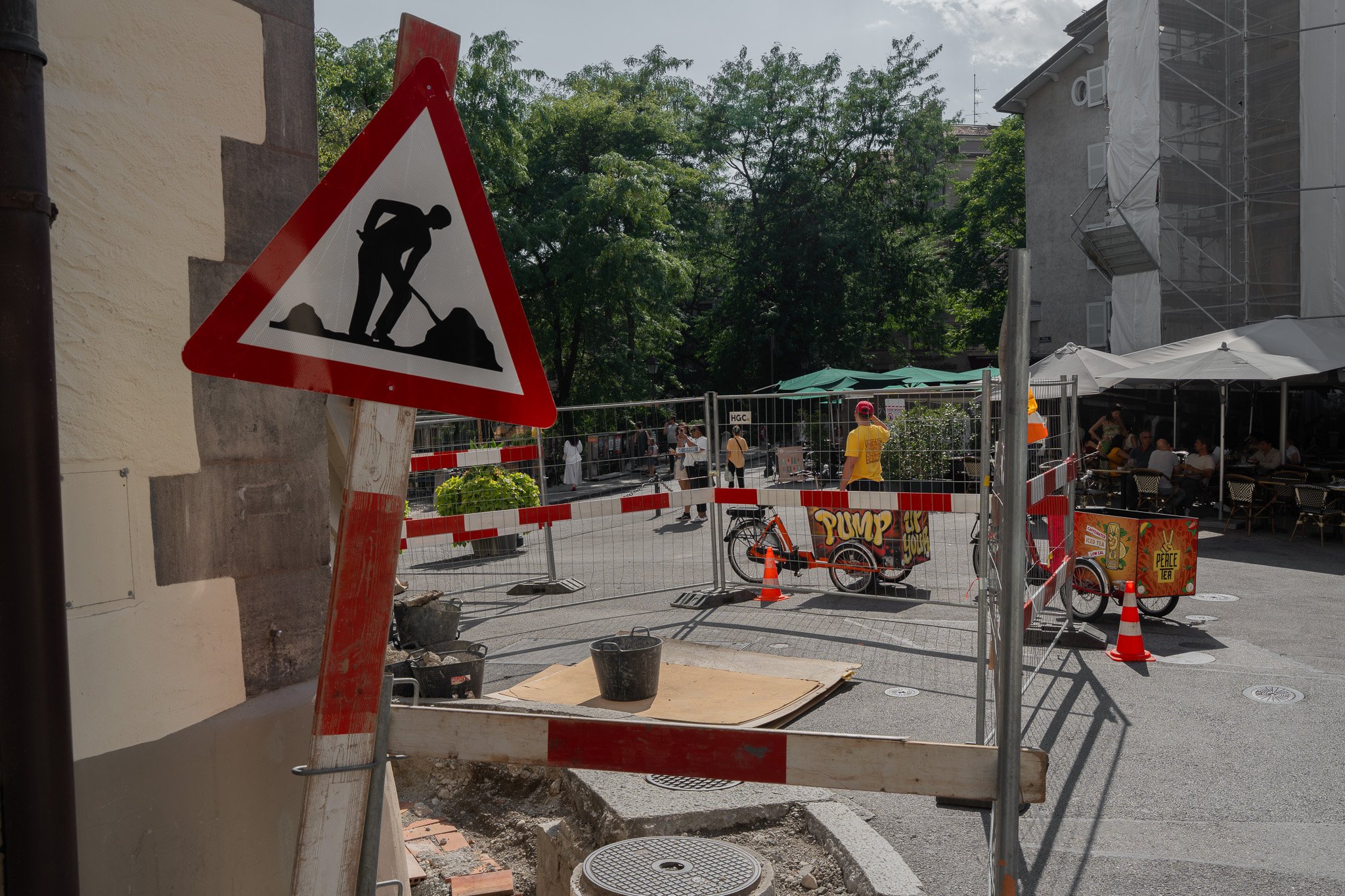 Le panneau de signalisation triangulaire montre une silhouette d'ouvrier avec une pelle dans un chantier de construction en milieu urbain, avec des arbres, des bâtiments, et des personnes assises à une terrasse à proximité.