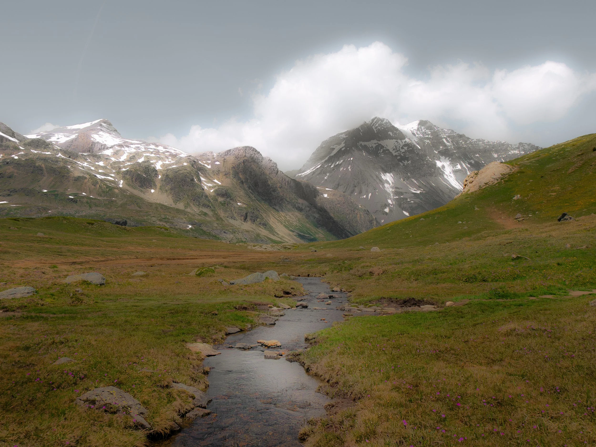 Paysage de montagnes rocheuses avec des sommets enneigés, une vallée verdoyante avec une rivière sinueuse au premier plan, arbres dispersés et un ciel partiellement nuageux.