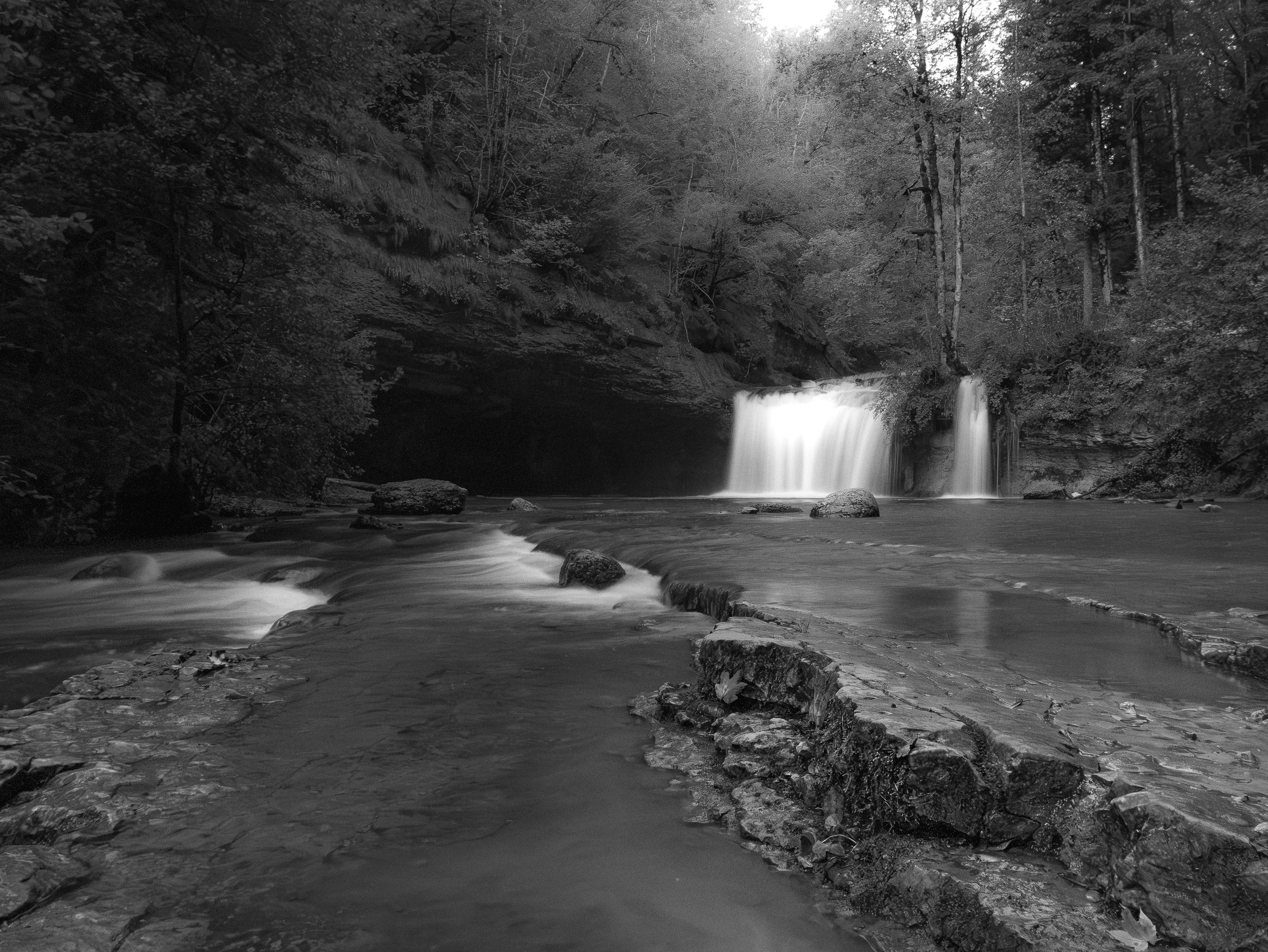 Une cascade coule dans une forêt dense avec des arbres, des rochers et de l'eau en mouvement dans un paysage naturel.