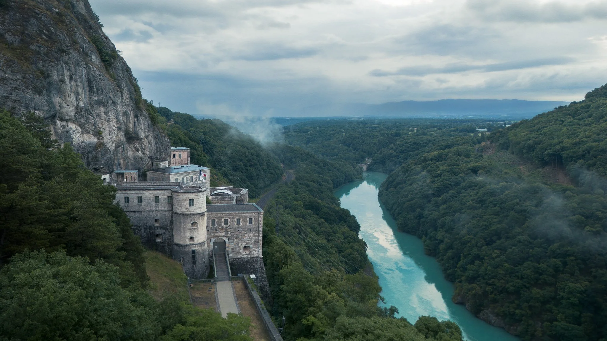 Une centrale hydroélectrique située dans une vallée verdoyante au bord d'une rivière turquoise sinueuse, entourée de collines boisées et de montagnes en arrière-plan sous un ciel nuageux.