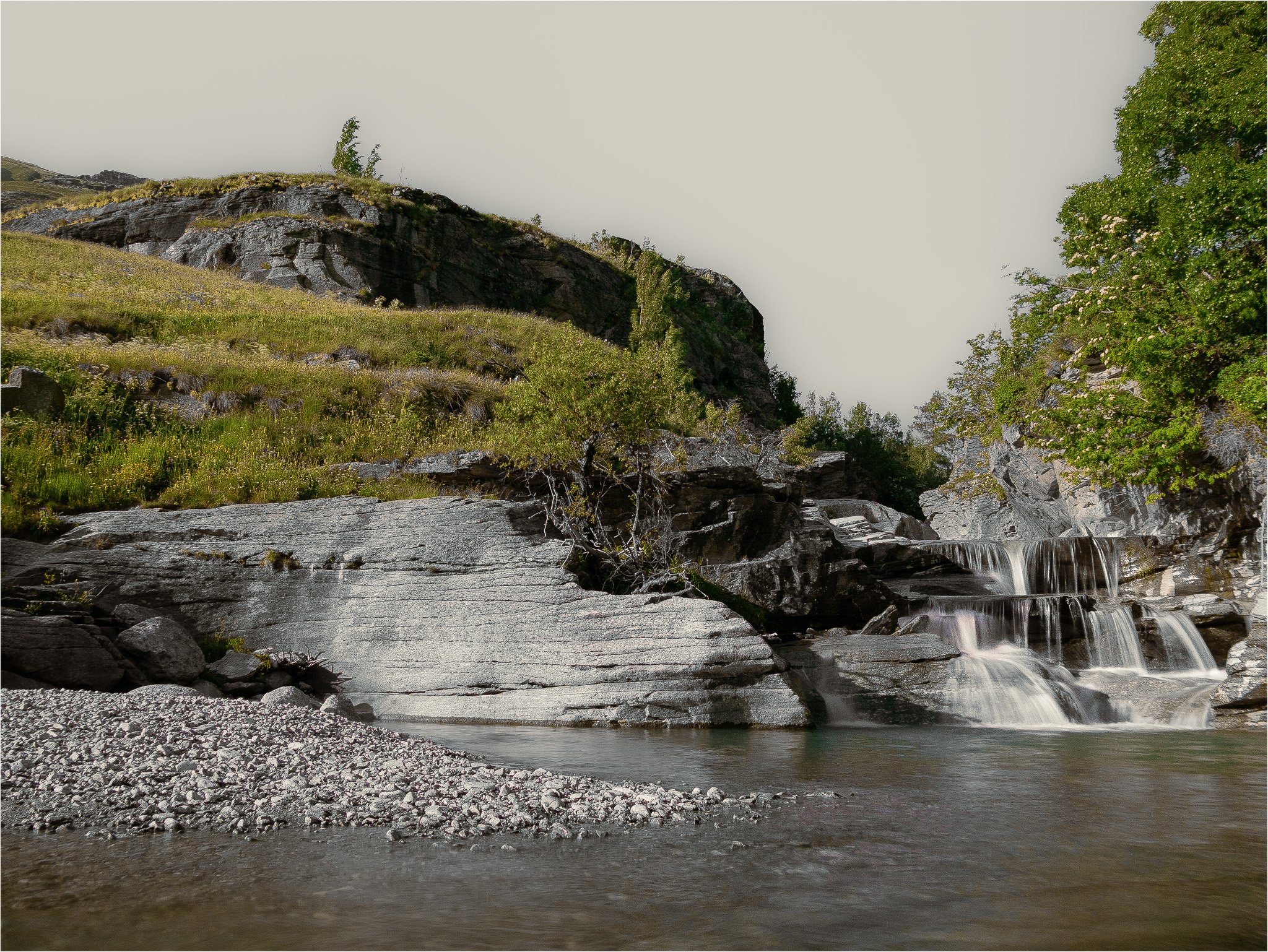 Cascade d'une rivière traversant une zone rocheuse entourée d'arbres et d'herbes verts, avec une falaise en arrière-plan et un ciel clair.