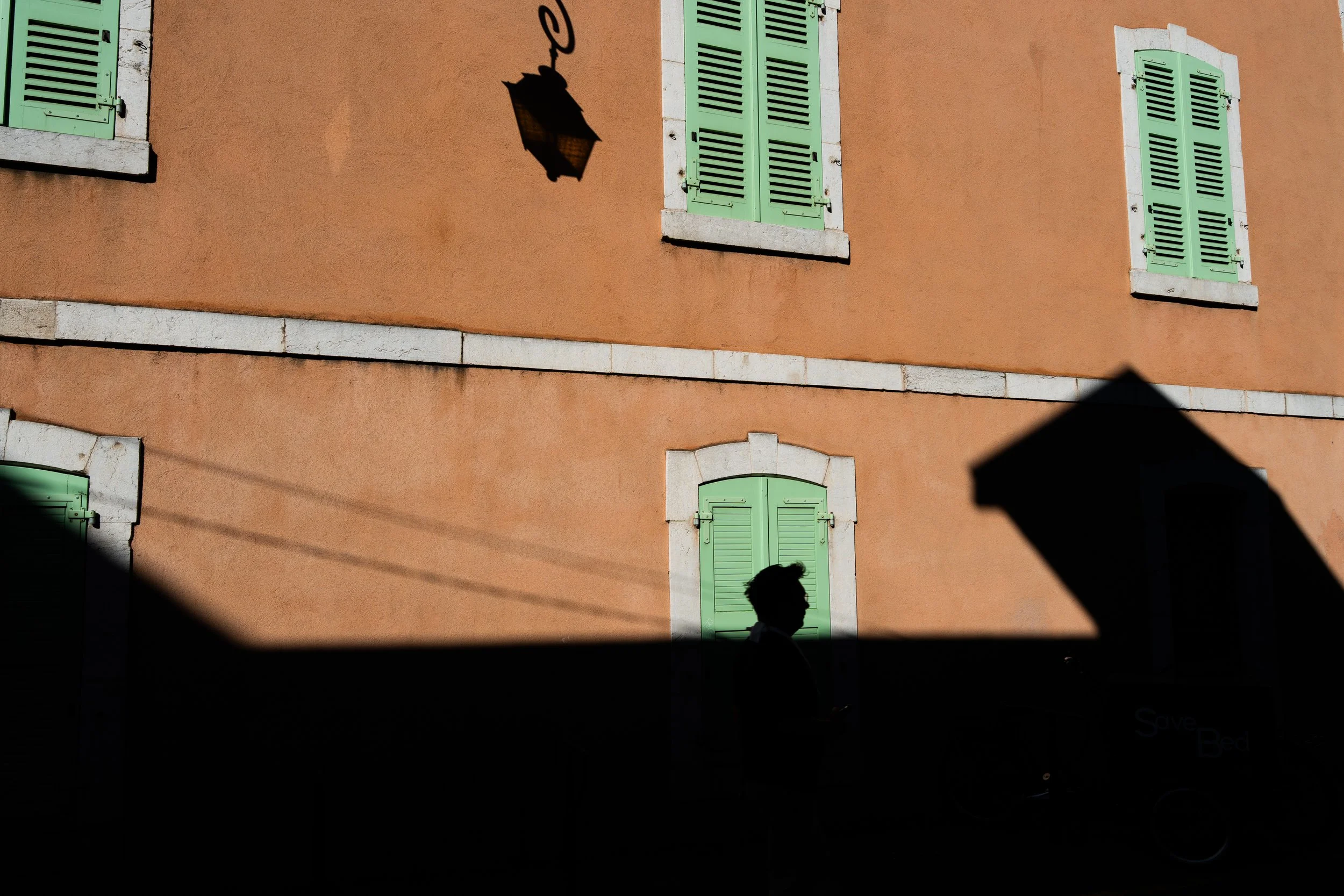 Une façade de bâtiment à couleur orange avec des fenêtres à volets vert pastel et encadrements en pierre blanche. La photo inclut des silhouettes de personnes en premier plan en contre-jour, ainsi qu'une ombre d'une lanterne sur le mur.