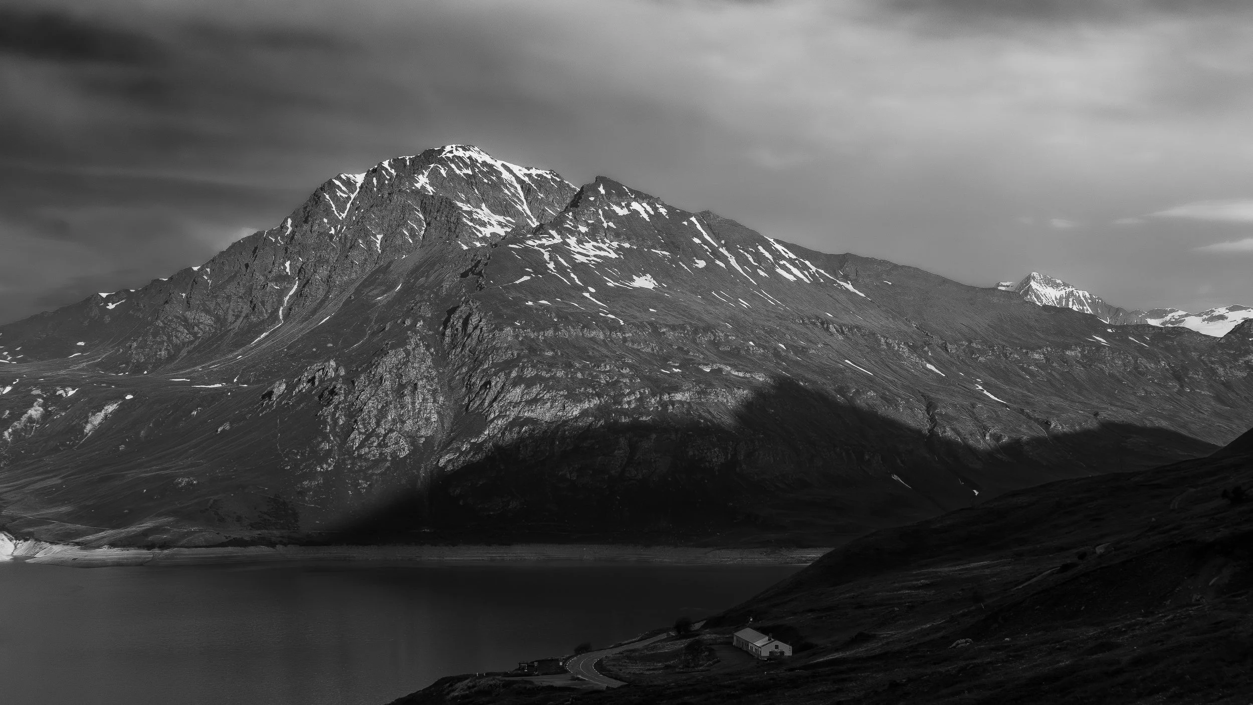 Une montagne enneigée surplombe un lac calme, avec un bâtiment isolé et une route serpentant dans le paysage. Le ciel est nuageux.