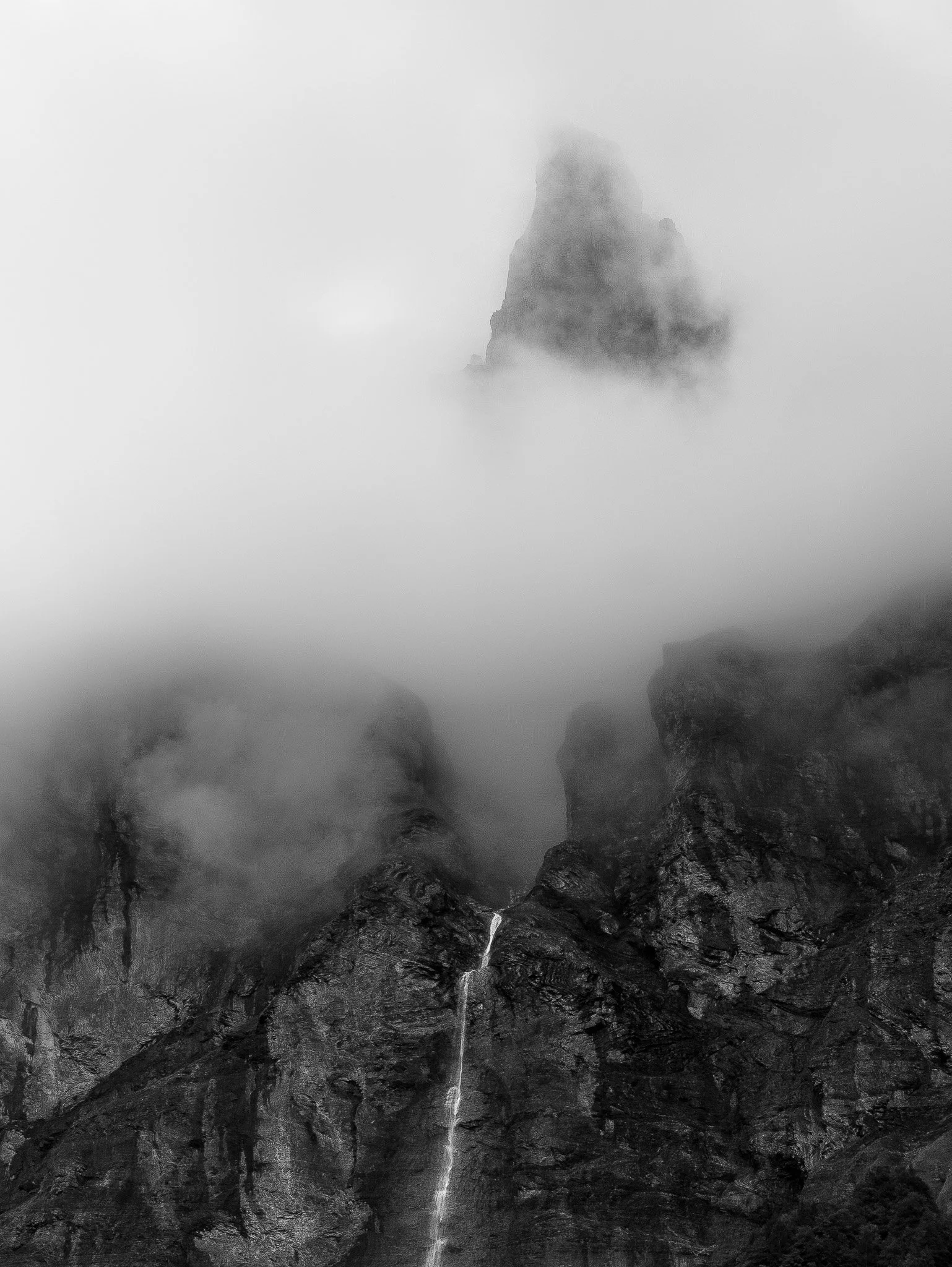 Une cascade tombe d'une montagne enneigée, entourée de rochers noirs, avec le brouillard et des nuages dans le ciel.