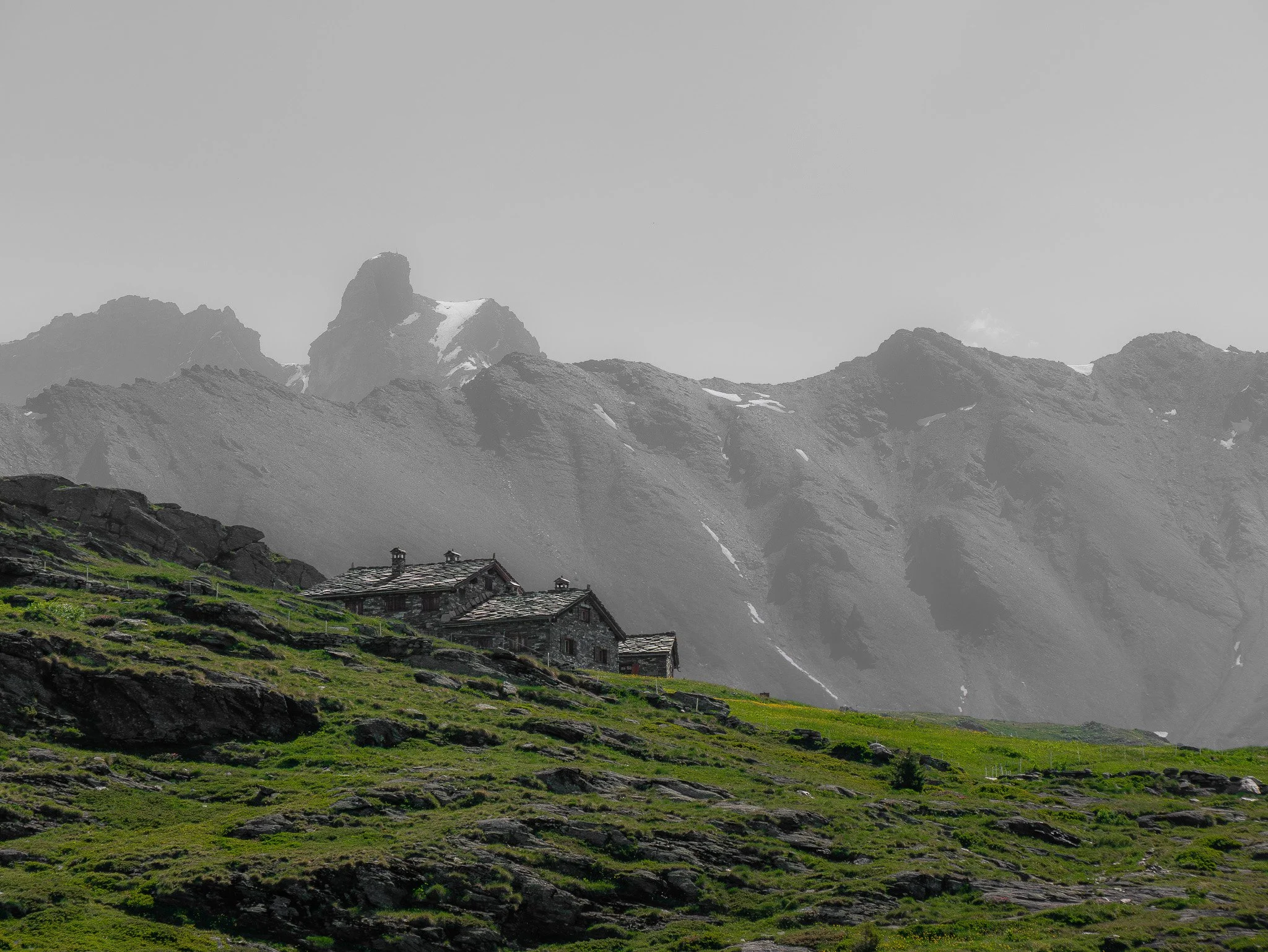 Montagnes enneigées en arrière-plan avec un village de cabanes en bois au premier plan, sur une pente herbeuse avec quelques rochers.