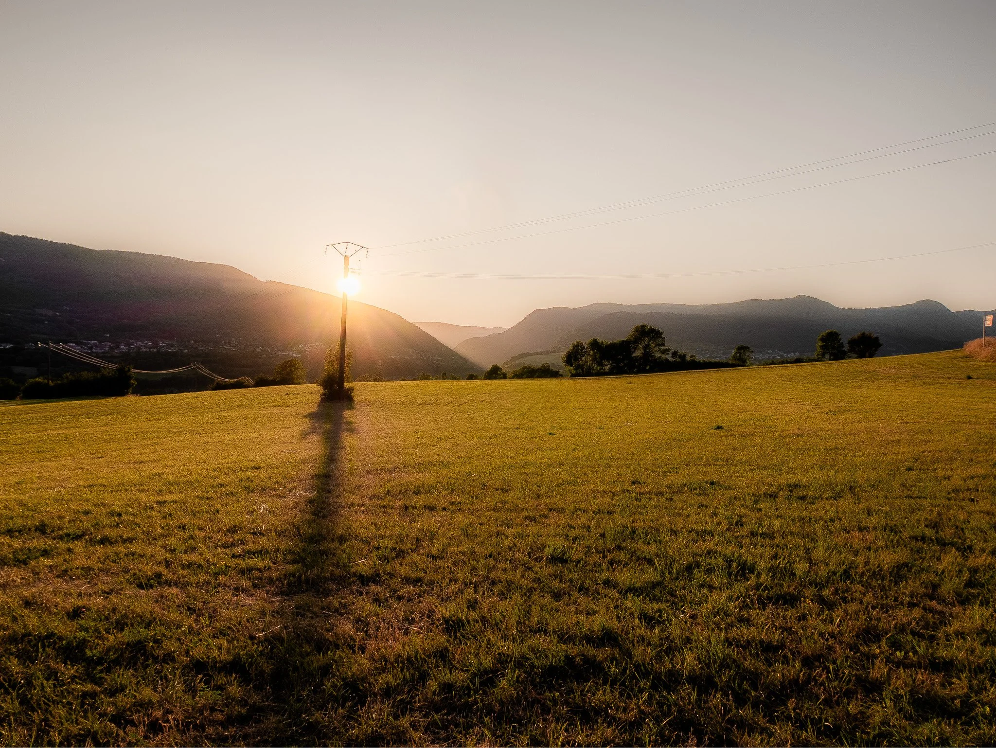 Paysage de campagne avec des collines et des arbres, un poteau électrique avec des fils, soleil derrière une colline, ciel clair