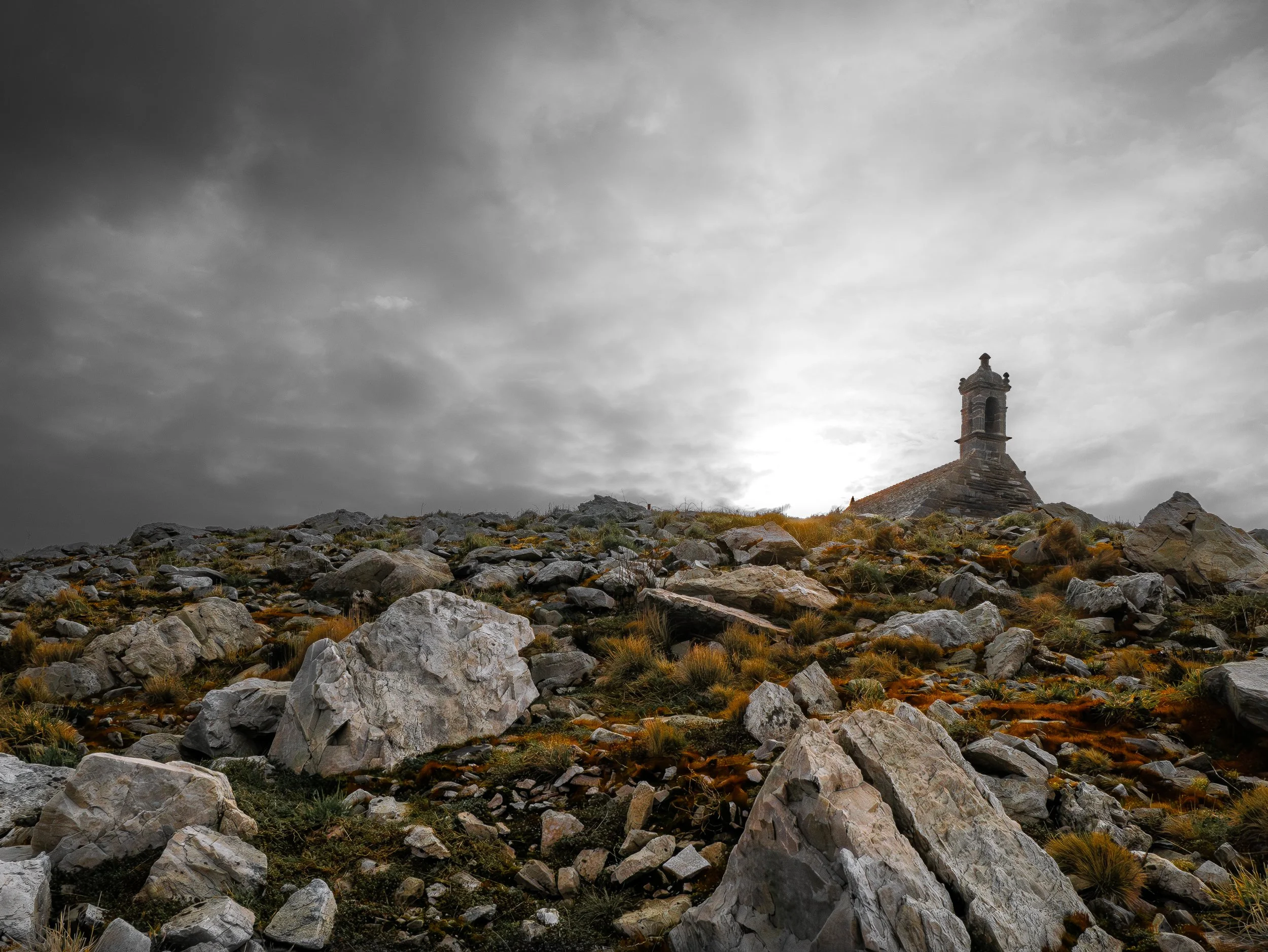 Une structure en pierre ressemblant à une petite chapelle ou un clocher situé sur une colline rocheuse, avec un ciel nuageux en arrière-plan.