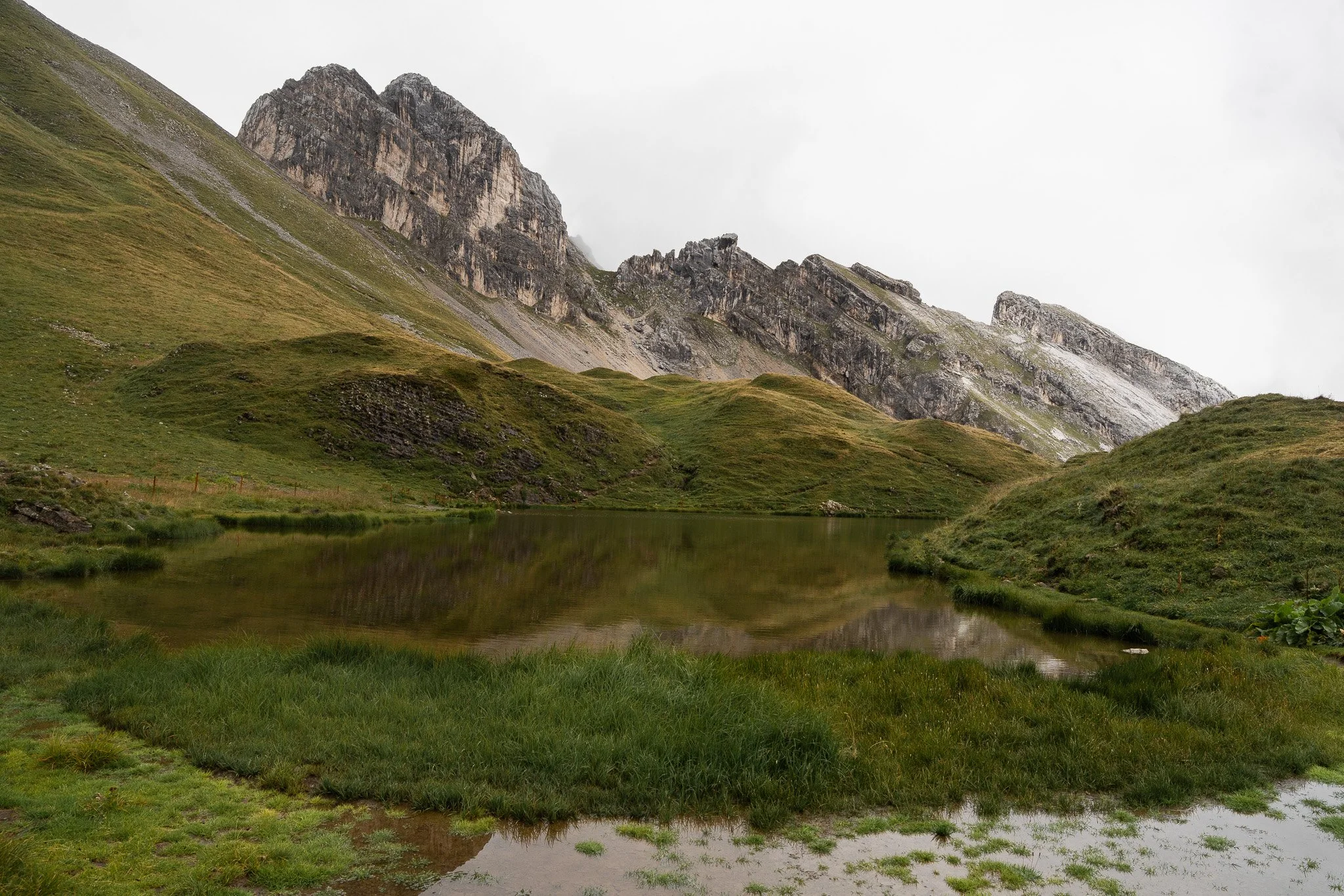 Montagne rocheuse avec des pentes recouvertes d'herbe, reflet dans un petit étang au premier plan, ciel nuageux