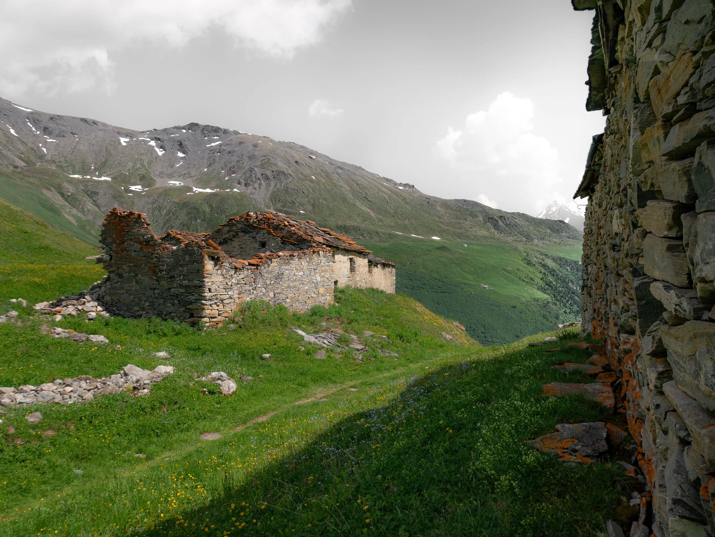 Paysage de montagnes avec des ruines en pierre et une structure rocheuse sur le côté droit. Le ciel est nuageux, la végétation est verte et abondante.
