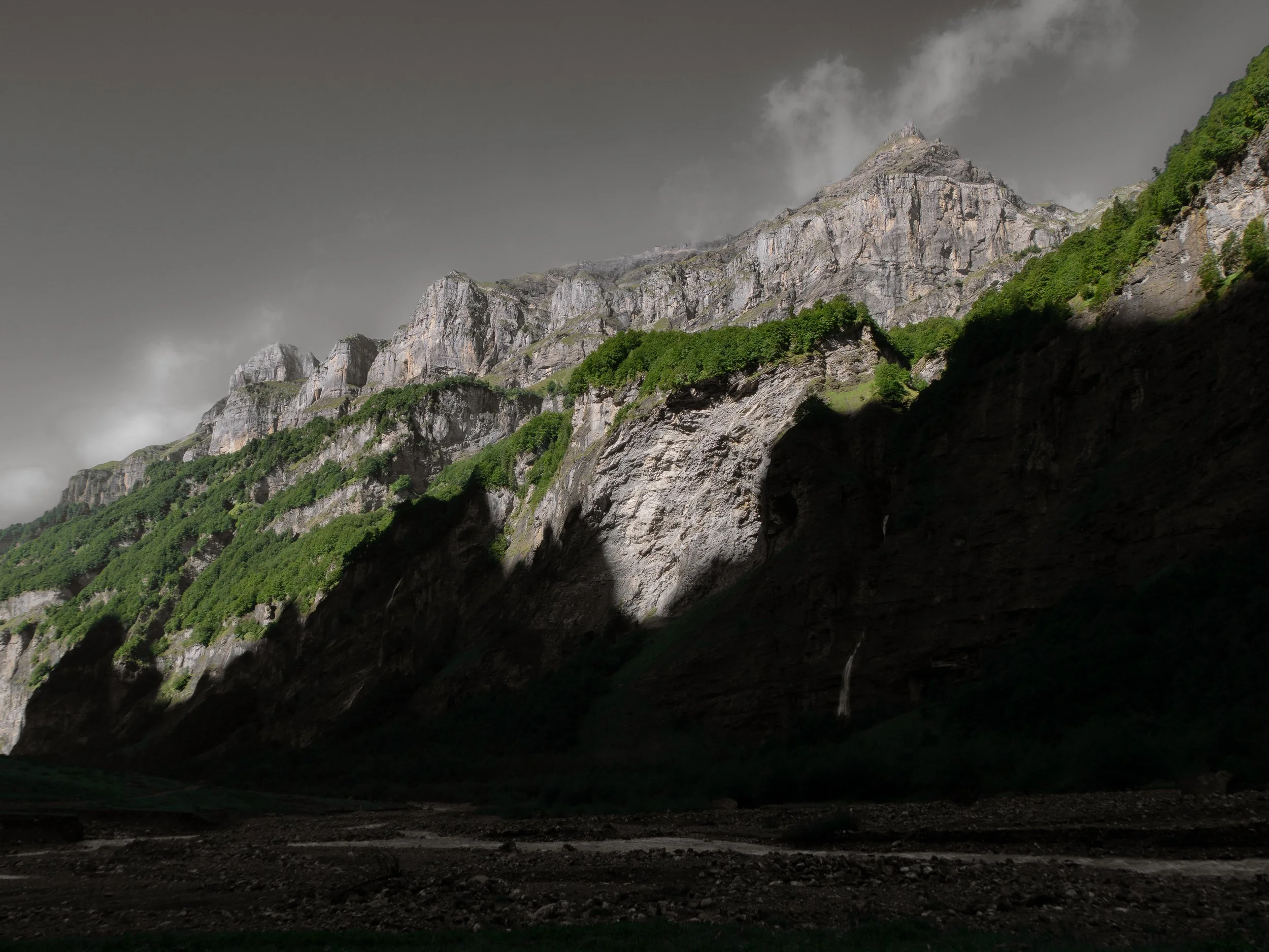 Montagnes rocheuses avec végétation verte, ciel gris orageux, et ombres profondes dans la vallée.