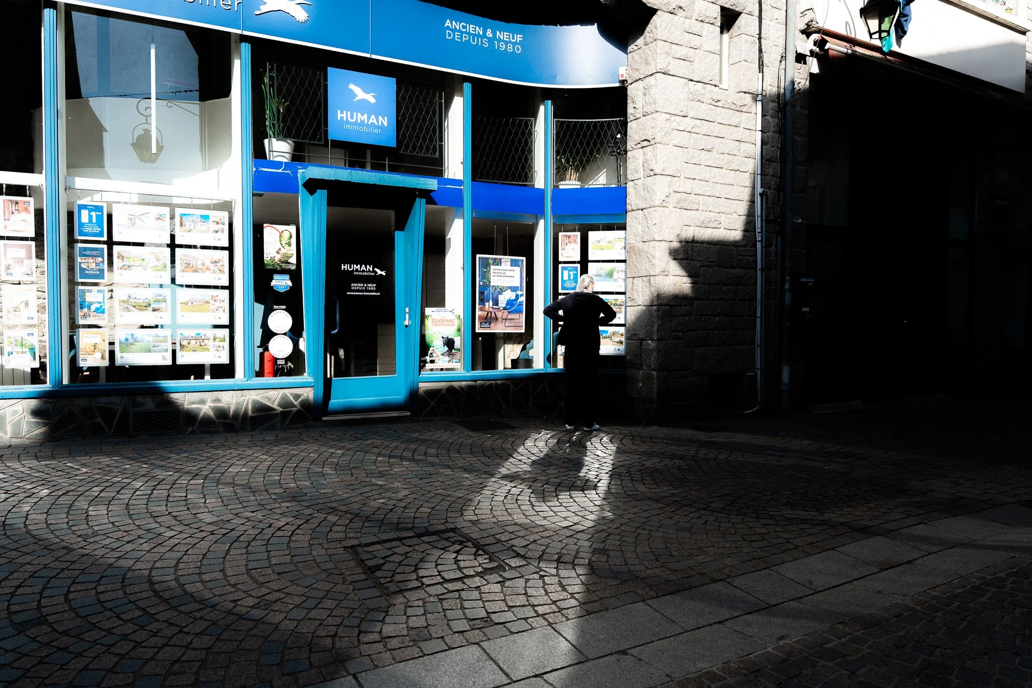 Une façade de boutique immobilière avec un grand panneau d'affichage, une porte en verre bleue et quelques affiches de propriétés à vendre ou louer. Un homme se tient devant la boutique, regardant le panneau.