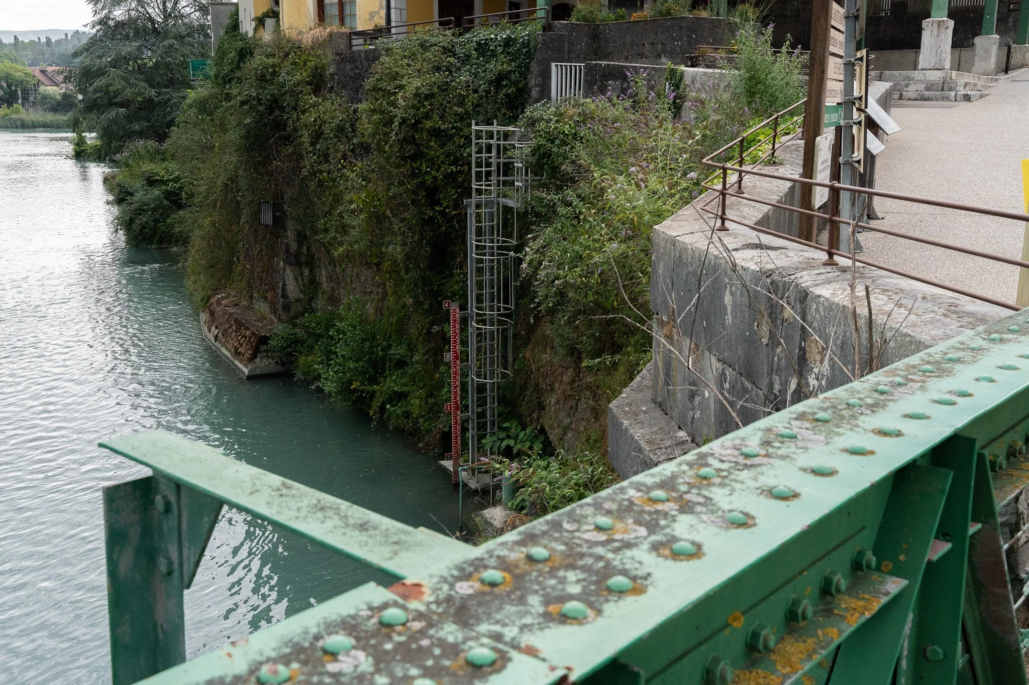 Vue d'une rivière ou d'un étang côté ville avec un pont en fer vert sur lequel on se trouve, vue vers la rive avec des végétations, des escaliers, une façade de bâtiment en jaune et du mobilier urbain.