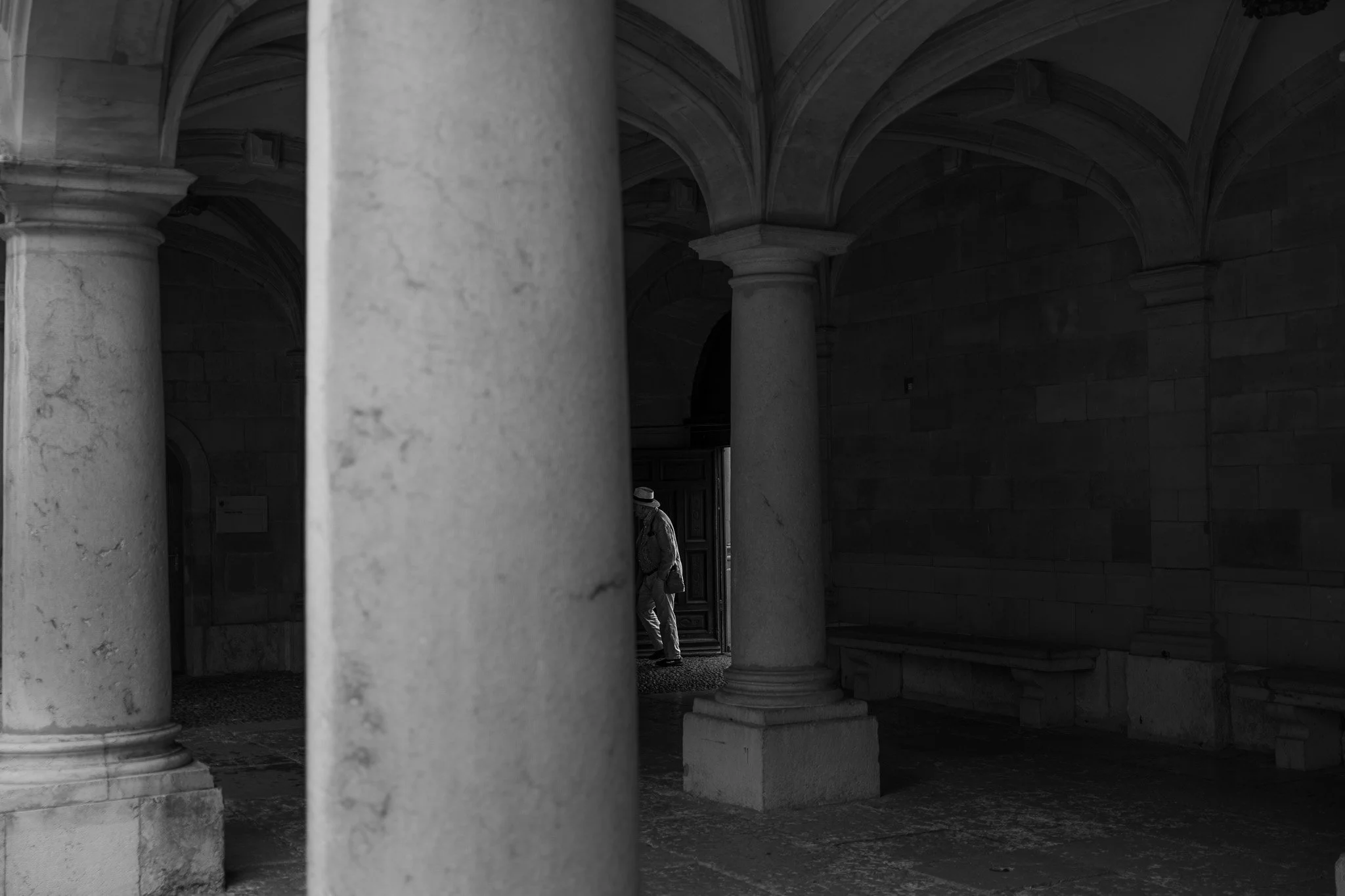 Photo en noir et blanc d'architecture avec de grandes colonnes en pierre et un plafond voûté, deux personnes visibles en arrière-plan près d'une porte.