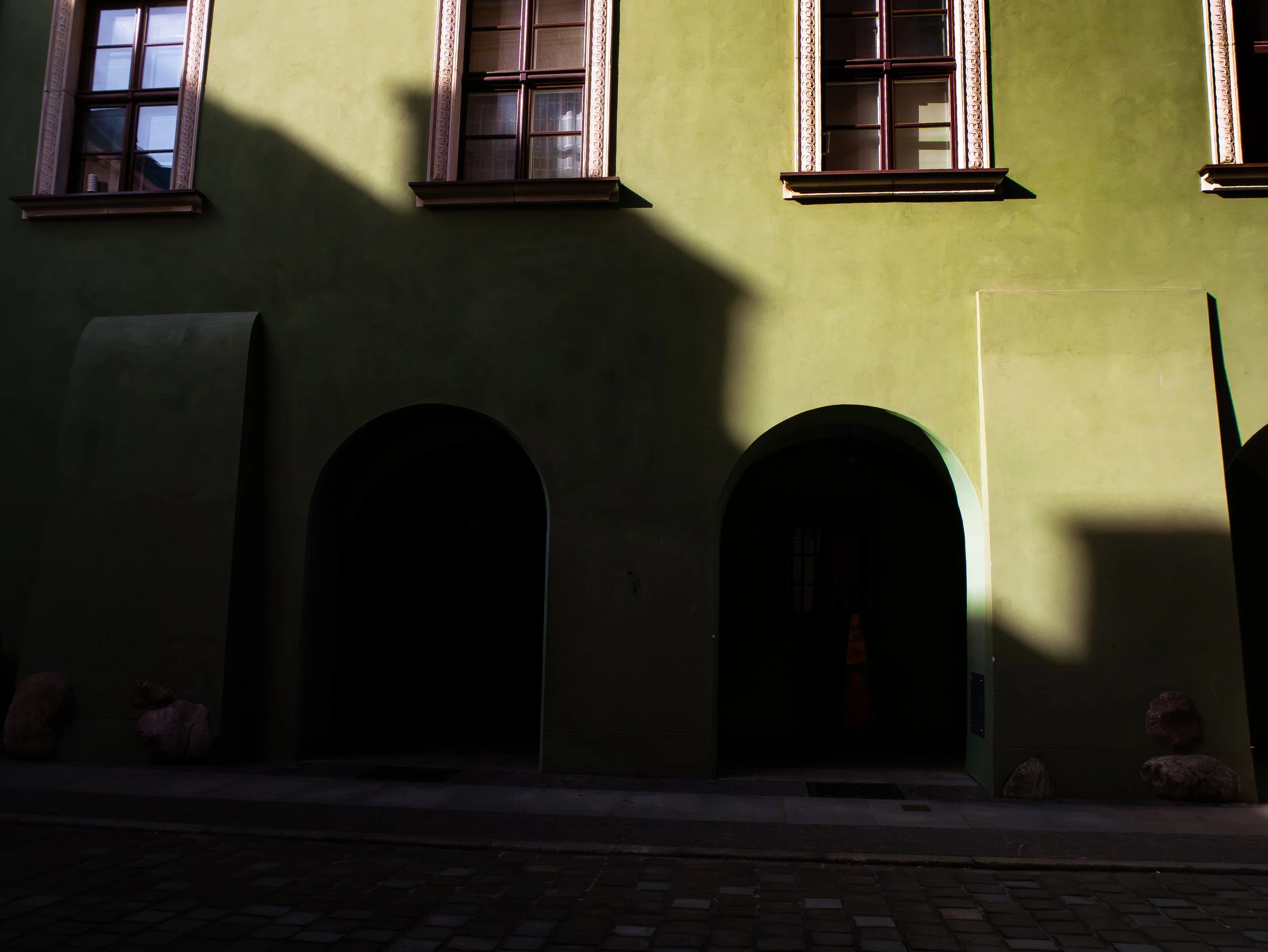 Façade d'un bâtiment vert avec trois fenêtres en haut. Ombre traversant la façade, partie inférieure sombre. Arches à la base du bâtiment.