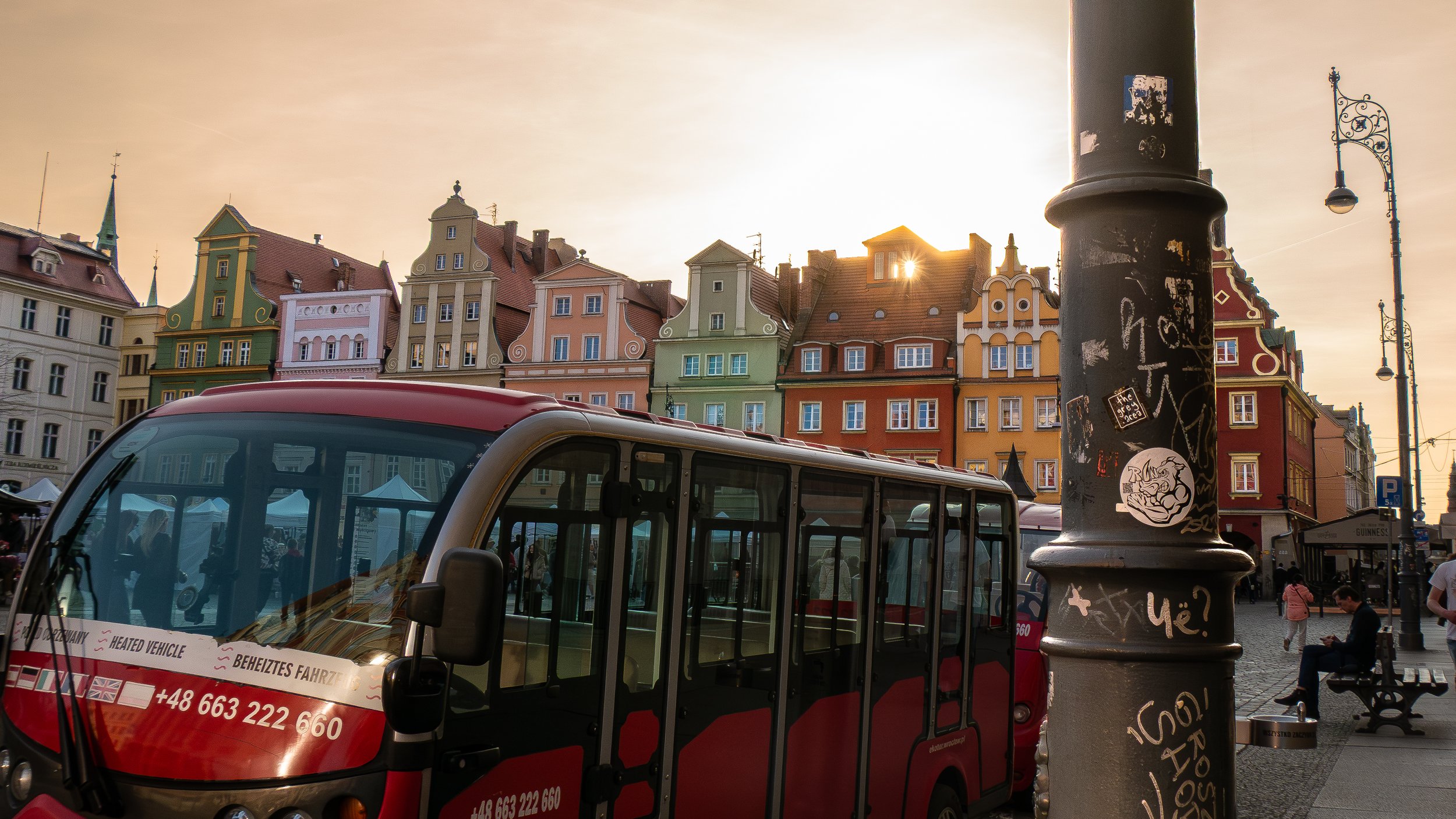 Une scène urbaine avec des bâtiments colorés à l'architecture traditionnelle, un bus électrique rouge, et un lampadaire noir graffiti. Le soleil derrière les bâtiments crée une ambiance chaleureuse.