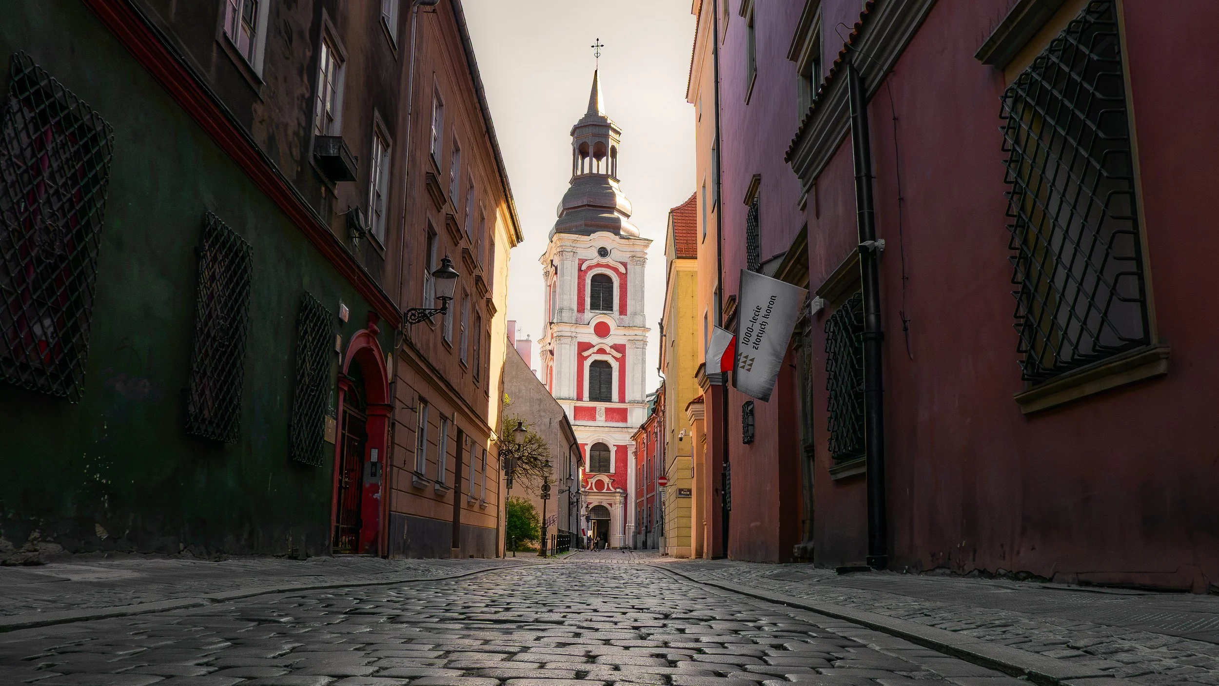 Une rue pavée bordée de bâtiments colorés menant à une église avec un clocher au fond, en fin d'après-midi avec une lumière dorée.