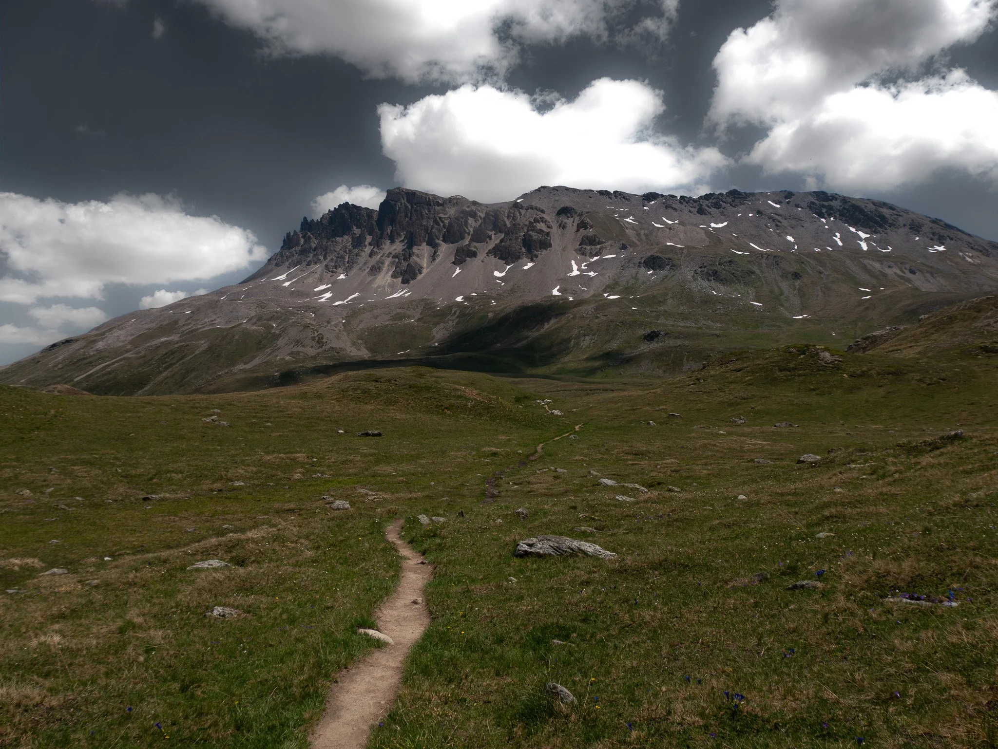 Une montagne avec des rochers escarpés, des zones enneigées et un ciel nuageux, vu d'une plaine verdoyante avec un sentier de terre.