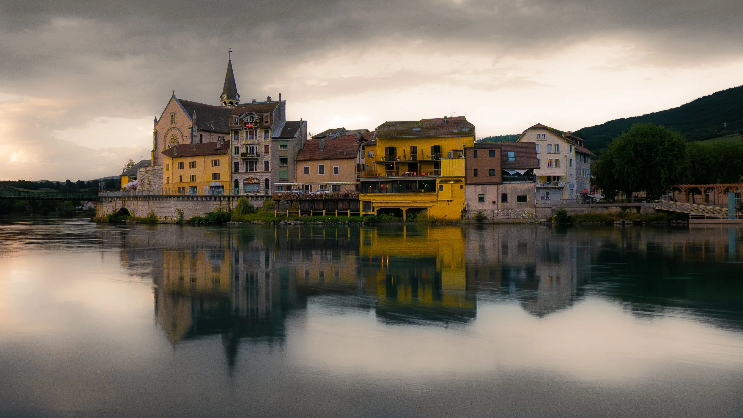 Un village coloré avec des bâtiments résidentiels et une grande église avec un clocher, reflété dans une rivière sous un ciel nuageux.